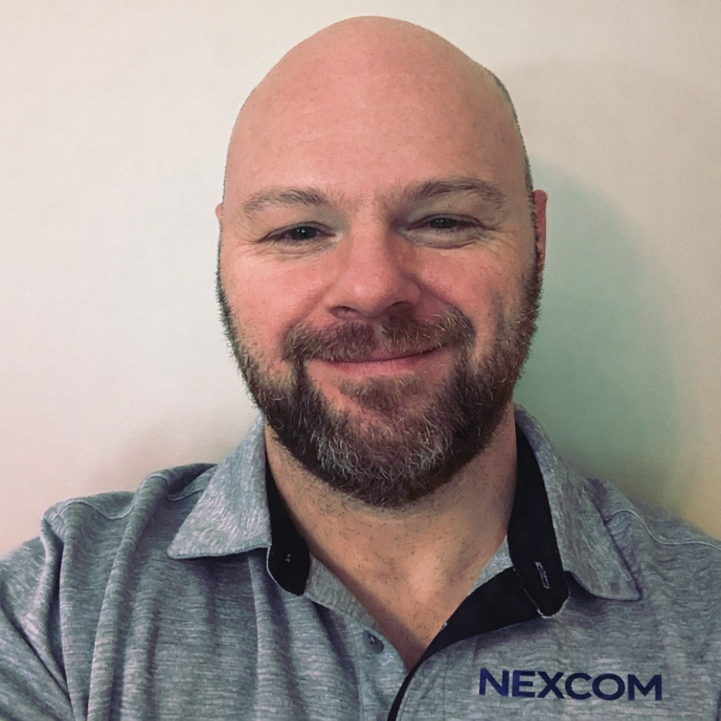 A smiling man with a beard and bald head wearing a gray polo shirt with NEXCOM logo, standing against a plain white wall.