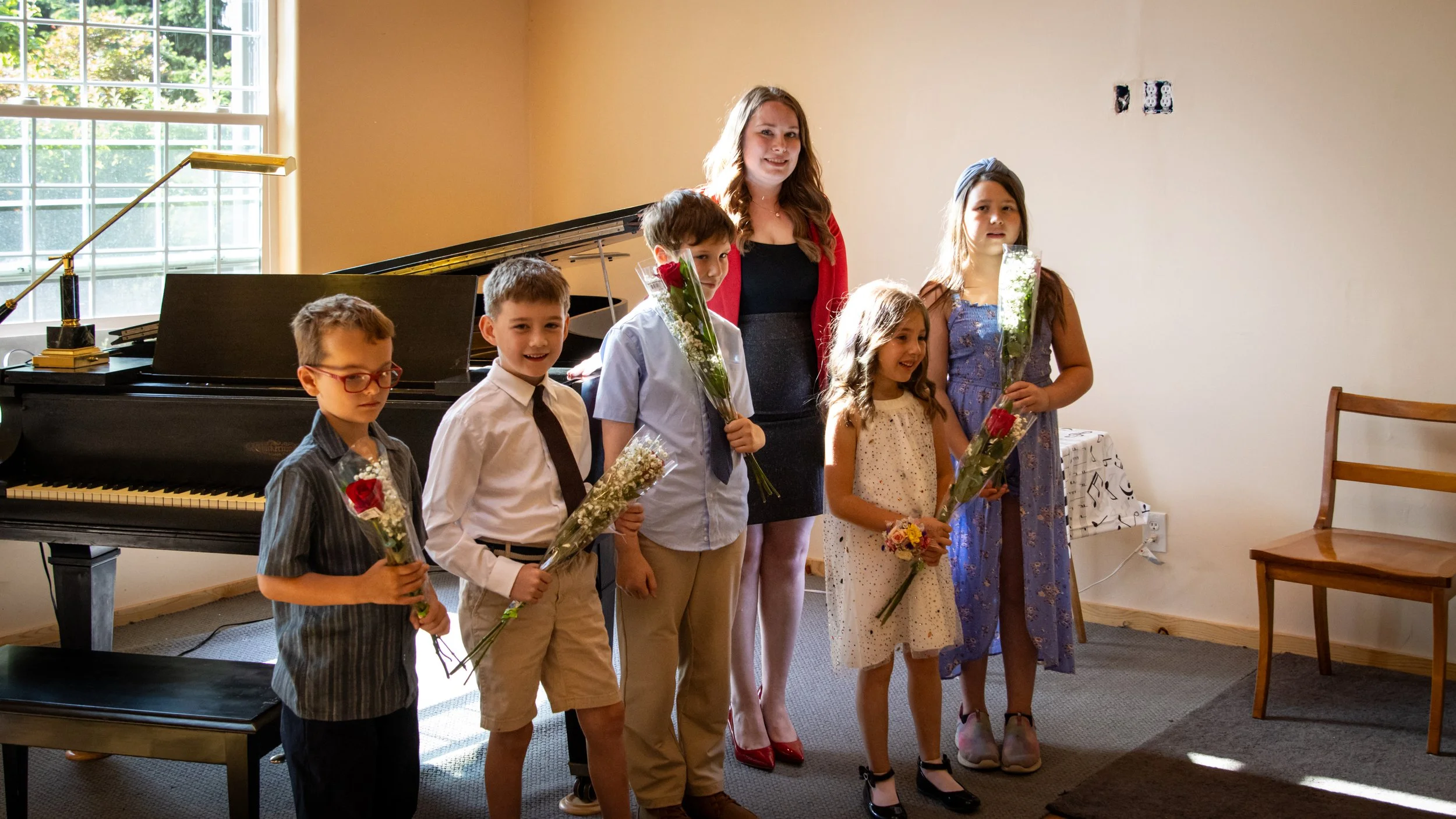 A woman and five children standing in a line indoors, holding flowers, with a grand piano in the background and a large window letting in natural light.