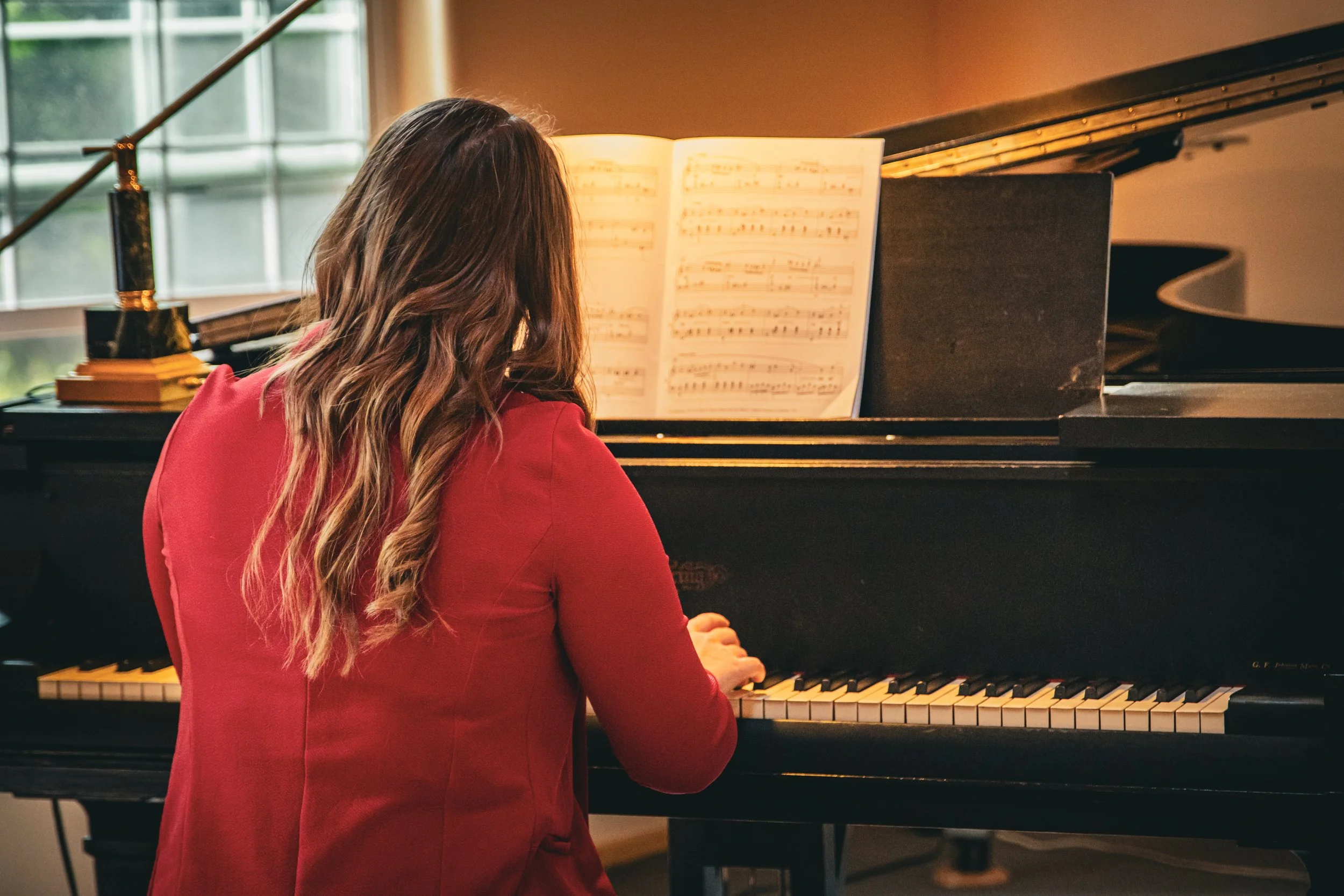 A woman with long wavy brown hair wearing a red blazer playing a black grand piano with an open sheet of music on the music stand.