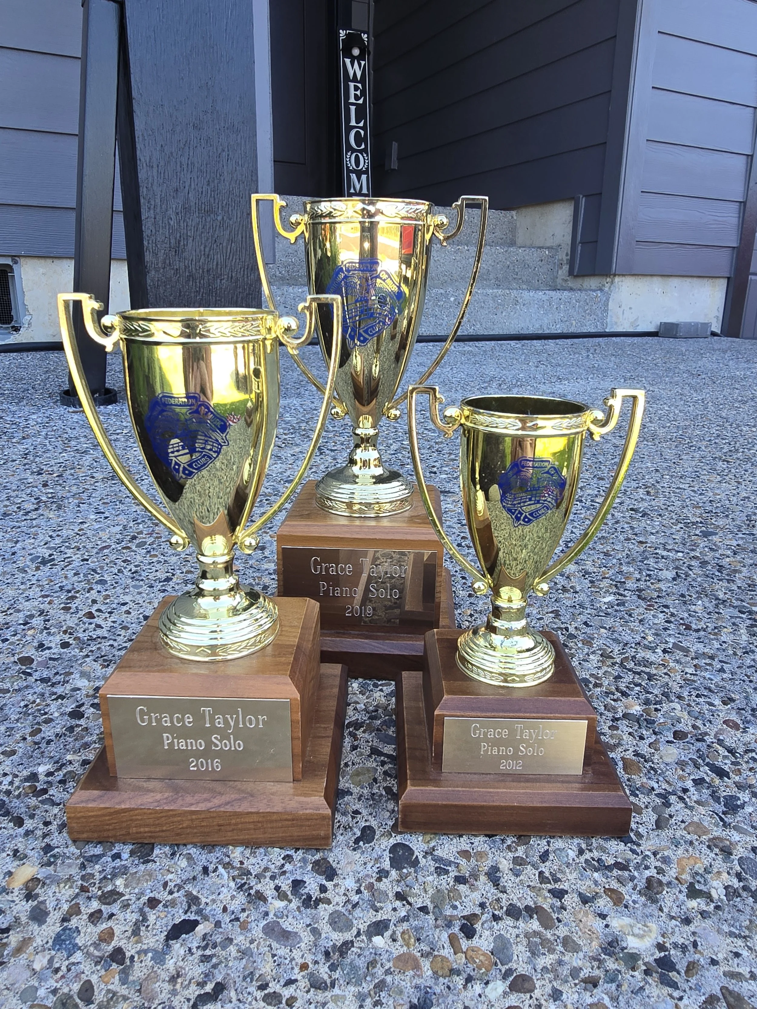 Three gold trophies on wooden bases with engraved plaques, displayed on a pebble-textured surface outside a building. The plaques read 'Grace Taylor Piano Solo' with years 2012, 2016, and 2019.