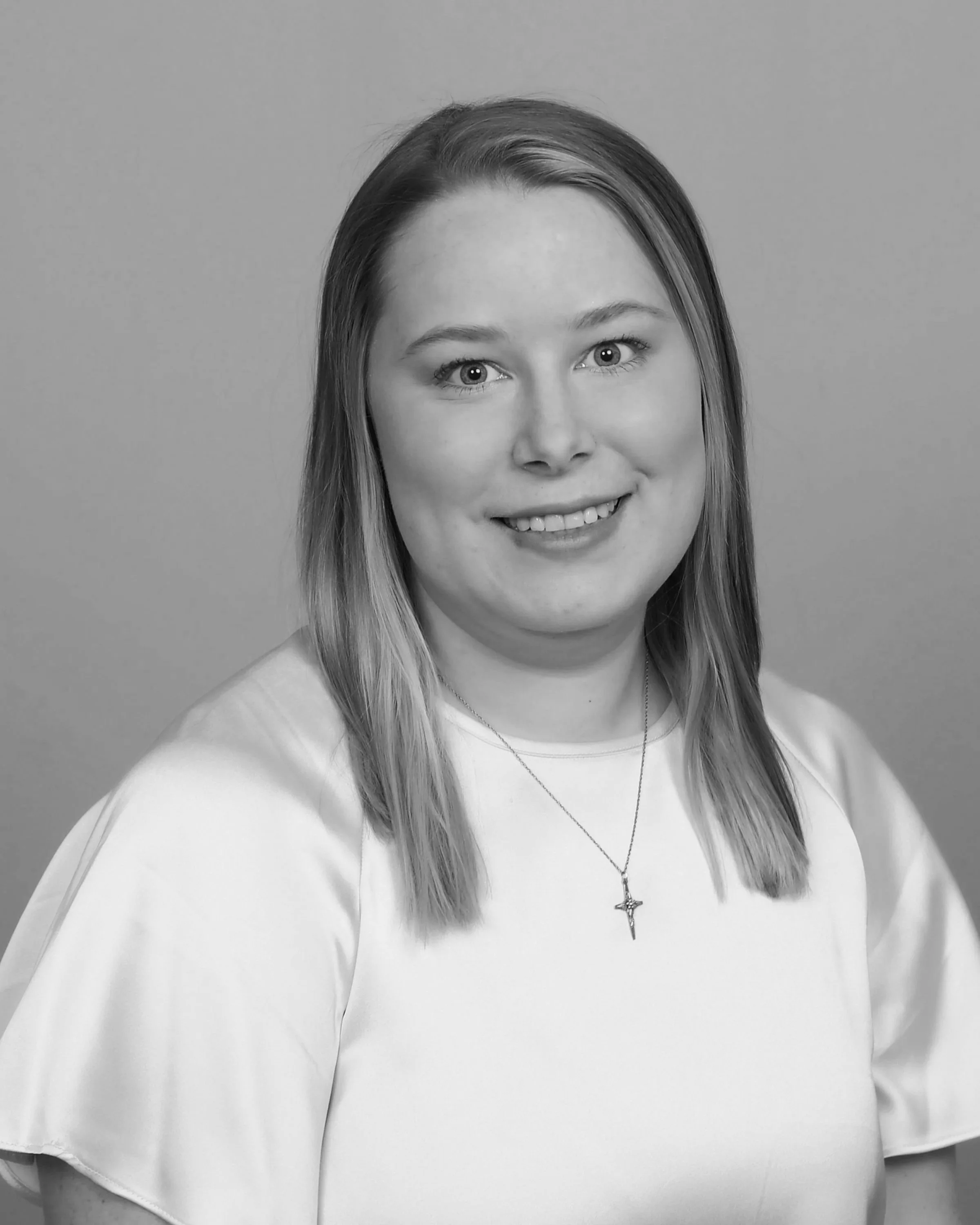 Black and white portrait of a smiling woman with shoulder-length hair, wearing a light-colored top and a necklace with a cross pendant, against a plain background.