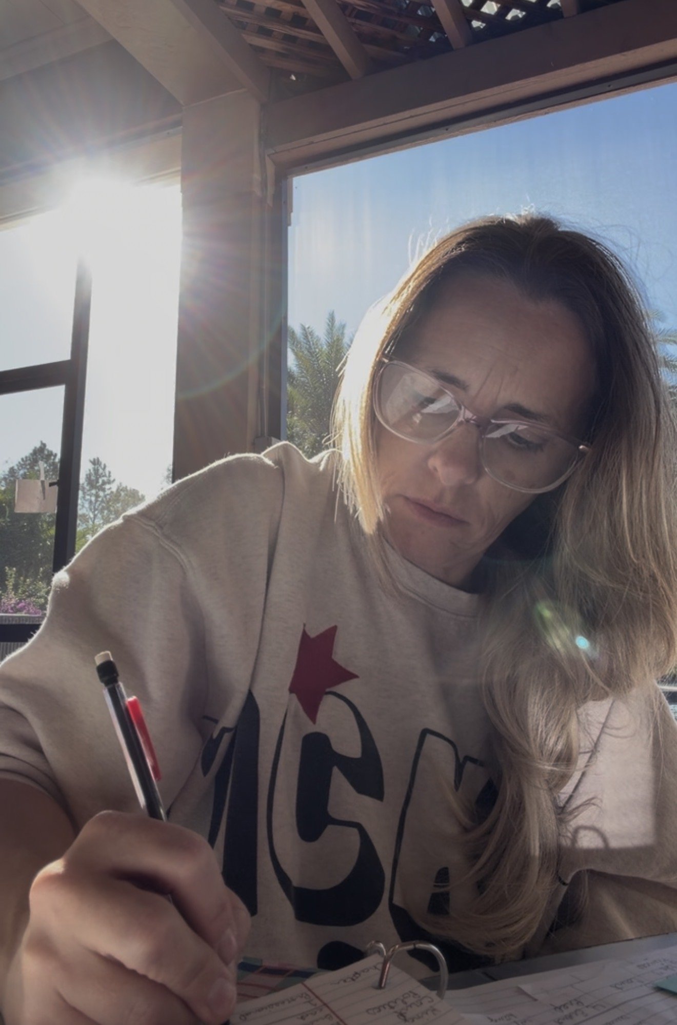 A woman with long blonde hair, wearing glasses and a cream-colored sweatshirt, is sitting at a desk and writing in a notebook. Sunlight is streaming through a large window behind her, with a view of trees and a clear blue sky outside.