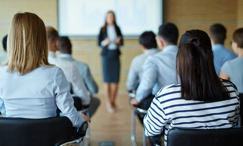 A woman in a blue patterned sweater is giving a presentation to a small group of people in a conference room, with a whiteboard behind her and a table with flowers, colorful markers, and mugs in front.