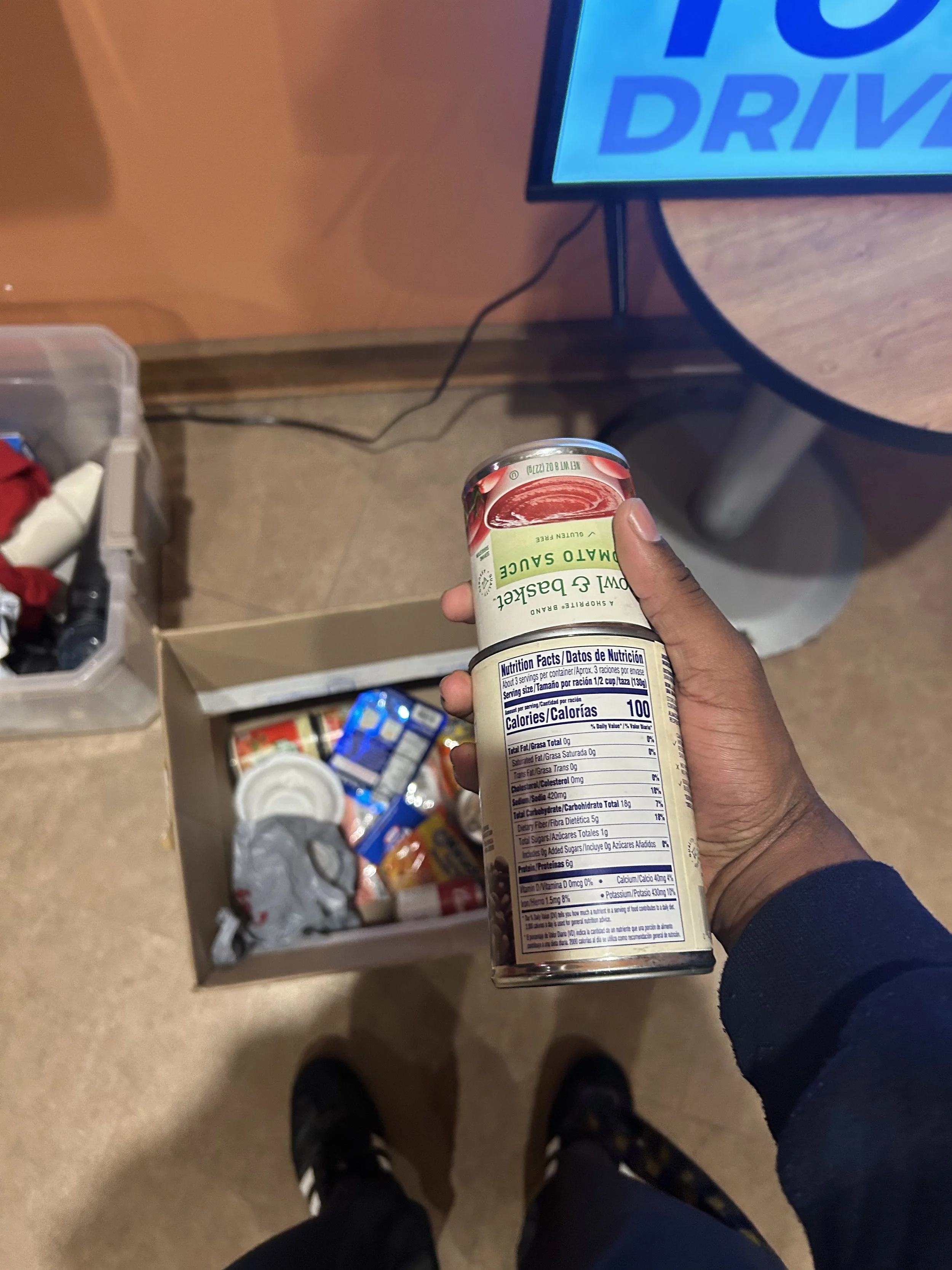 A person holding a canned food item over a box of various snacks and drinks. In the background, there is a table with a screen showing 'DRIVE' and some loose items on the floor.