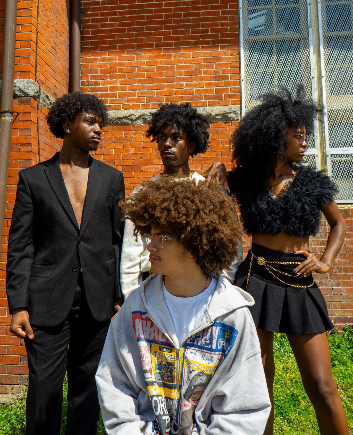 Four young adults with Afro hairstyles standing and sitting against a brick building with a metal fire escape. One person is wearing a black blazer, another in a white sweatshirt, a third in a black feathered crop top, and the fourth in an oversized graphic hoodie. They appear to be posing outdoors in daylight.