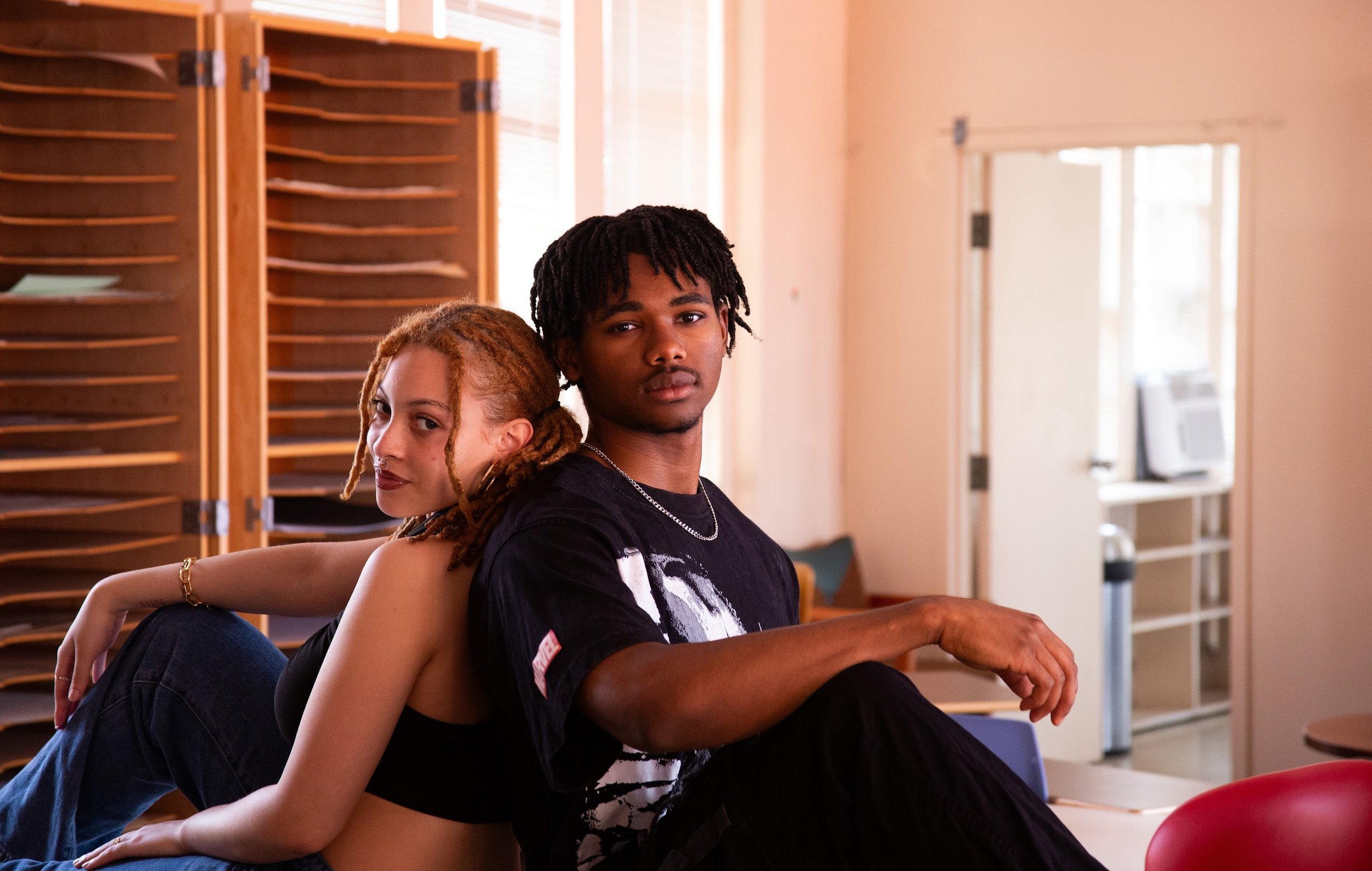 Two young individuals, a woman with blonde dreadlocks and a man with black dreadlocks, sit back-to-back in a room with a wooden cabinet, a window, and an air conditioner in the background.