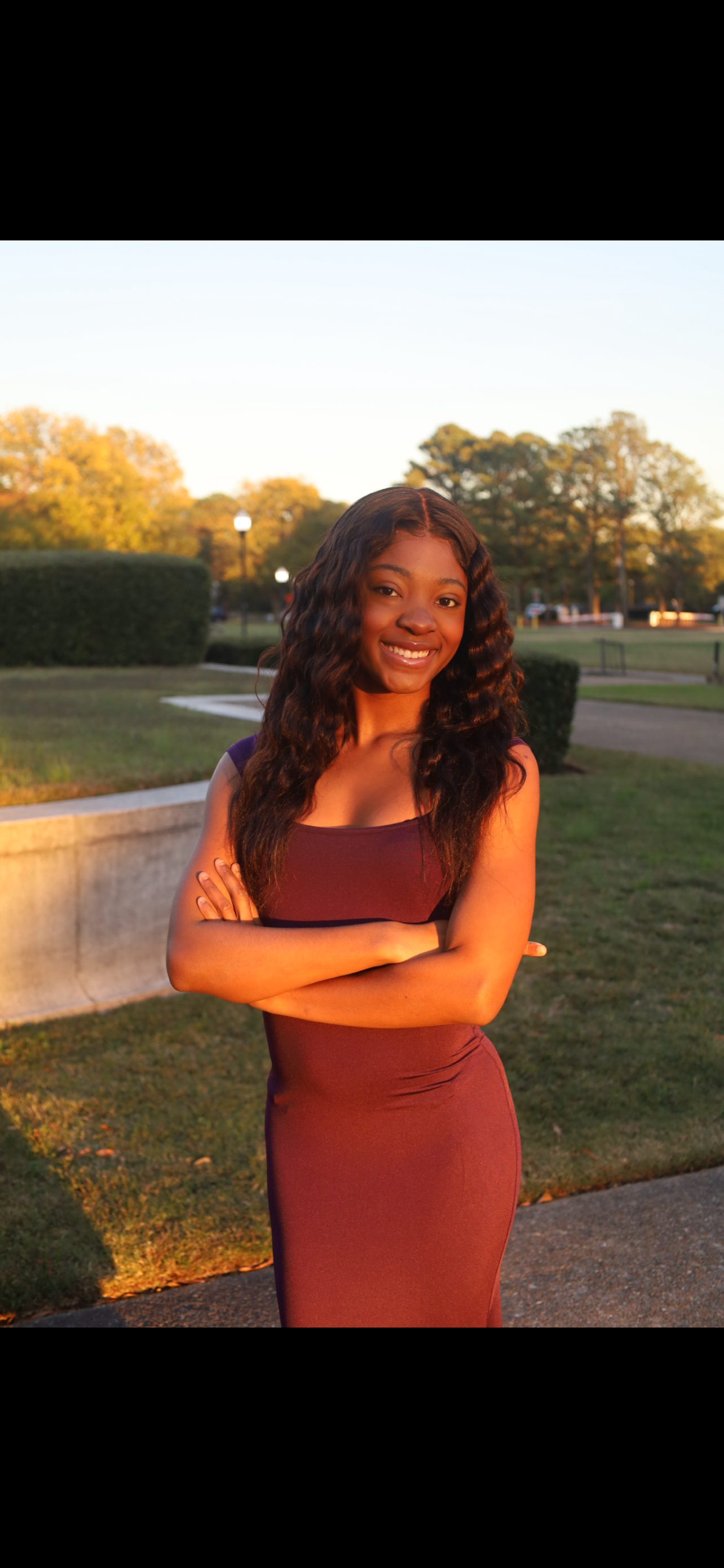 Young woman with long wavy dark hair smiling, arms crossed, wearing a sleeveless burgundy dress, standing outdoors during sunset in a park with trees, grass, pathway, and street lamps in the background.
