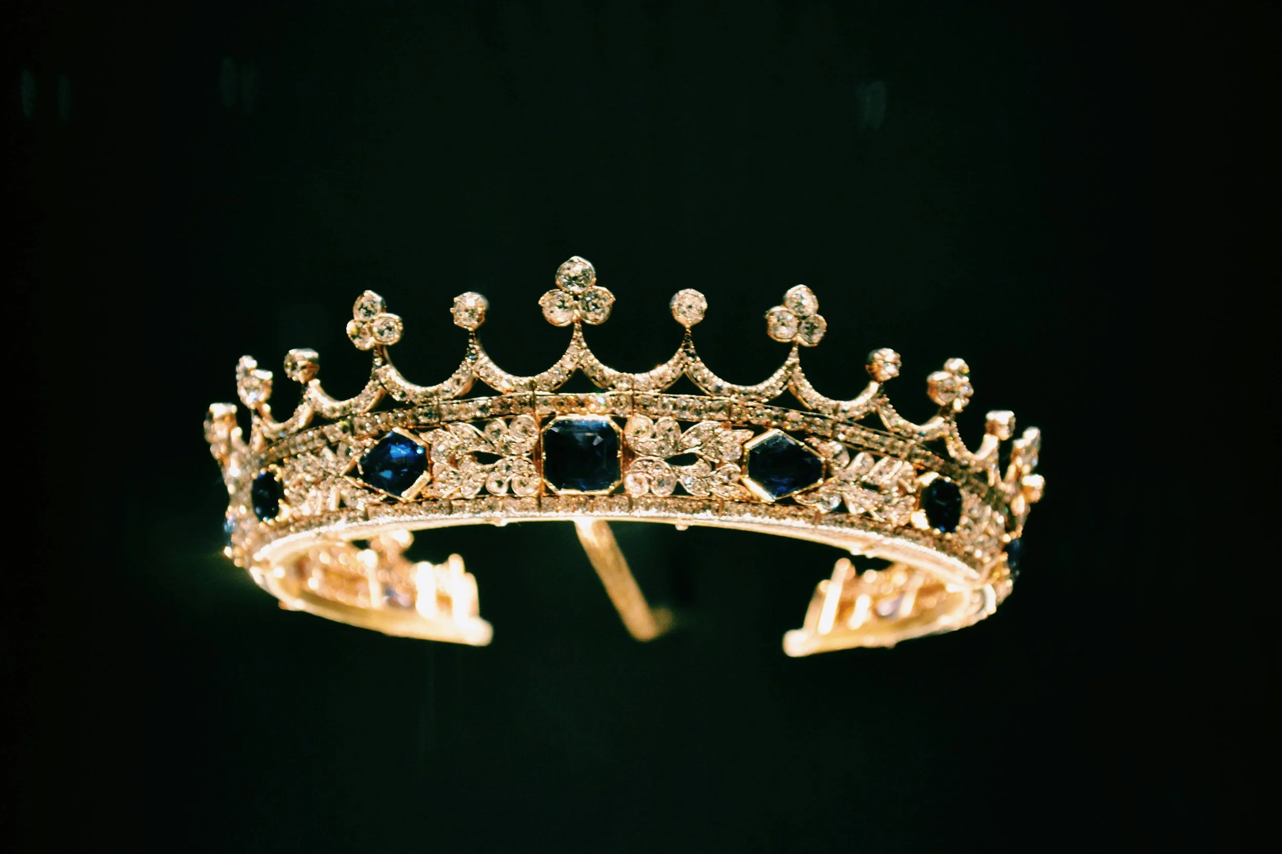 A gold crown adorned with blue and clear gemstones against a black background.