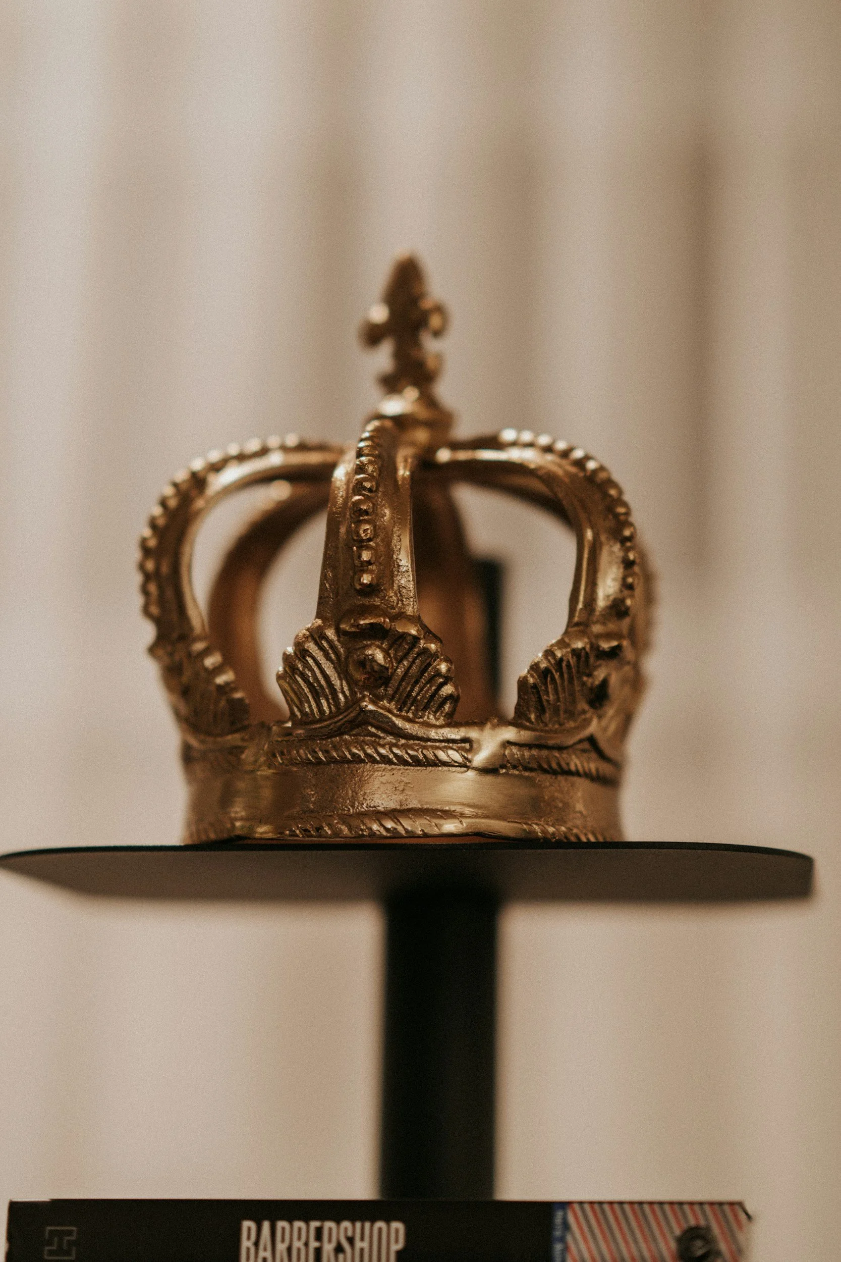 A small metallic golden crown sculpture on a round platform, placed on a stack of books or magazines.