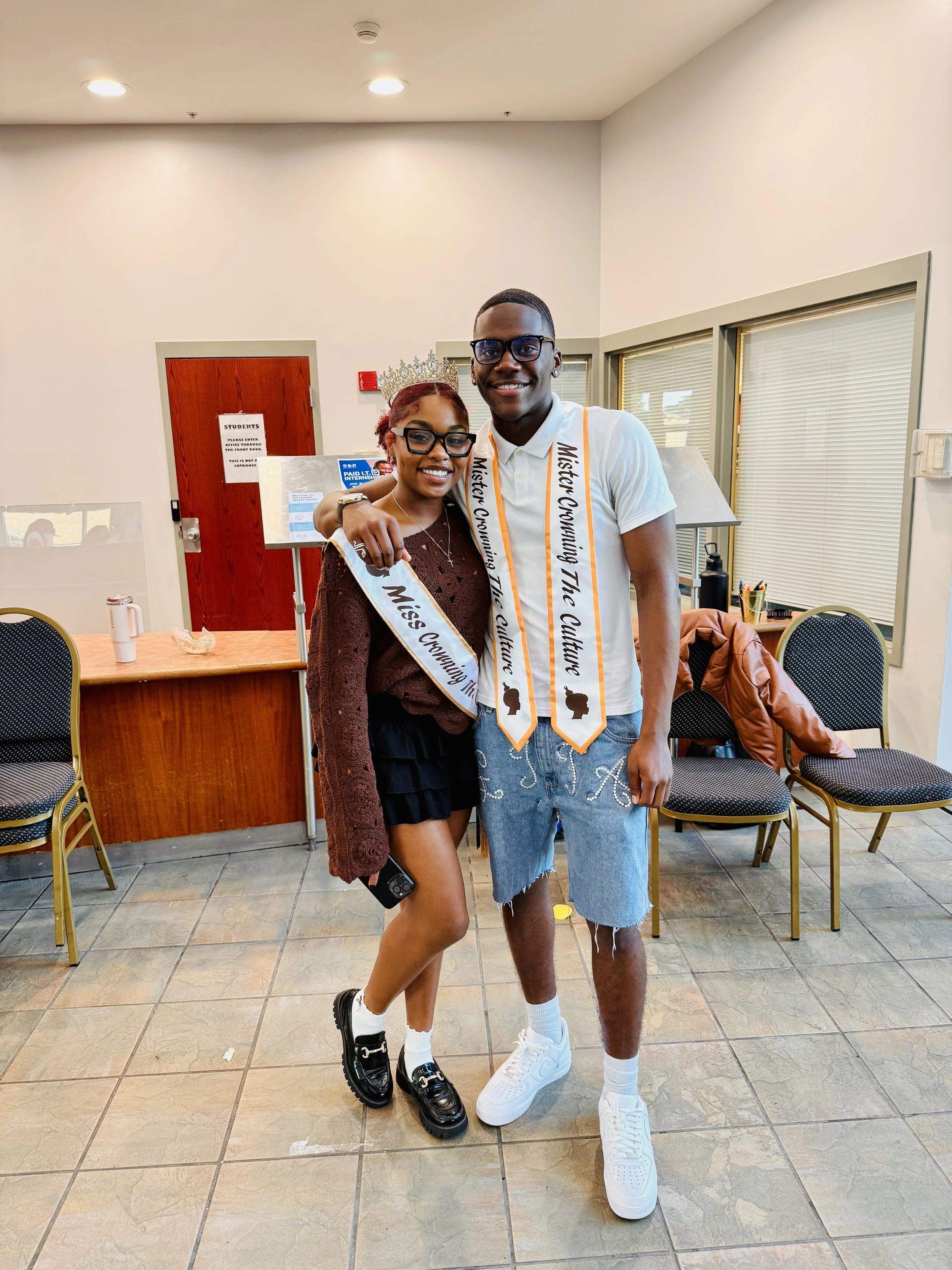 Two young adults smiling, one wearing a crown and sash that reads 'Miss Cunnning...' and the other wearing a sash that reads 'Mystery Crossing The Culture', posing in a room with chairs, window blinds, and a desk in the background.