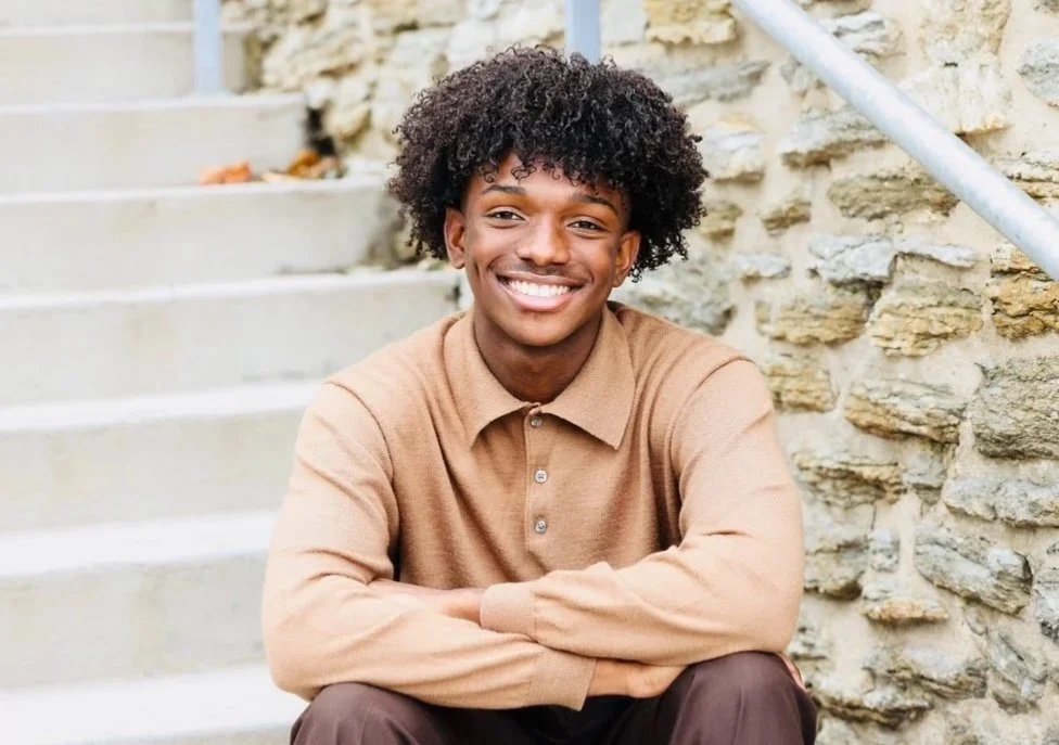 A young man smiling while sitting on steps outdoors near a stone wall.