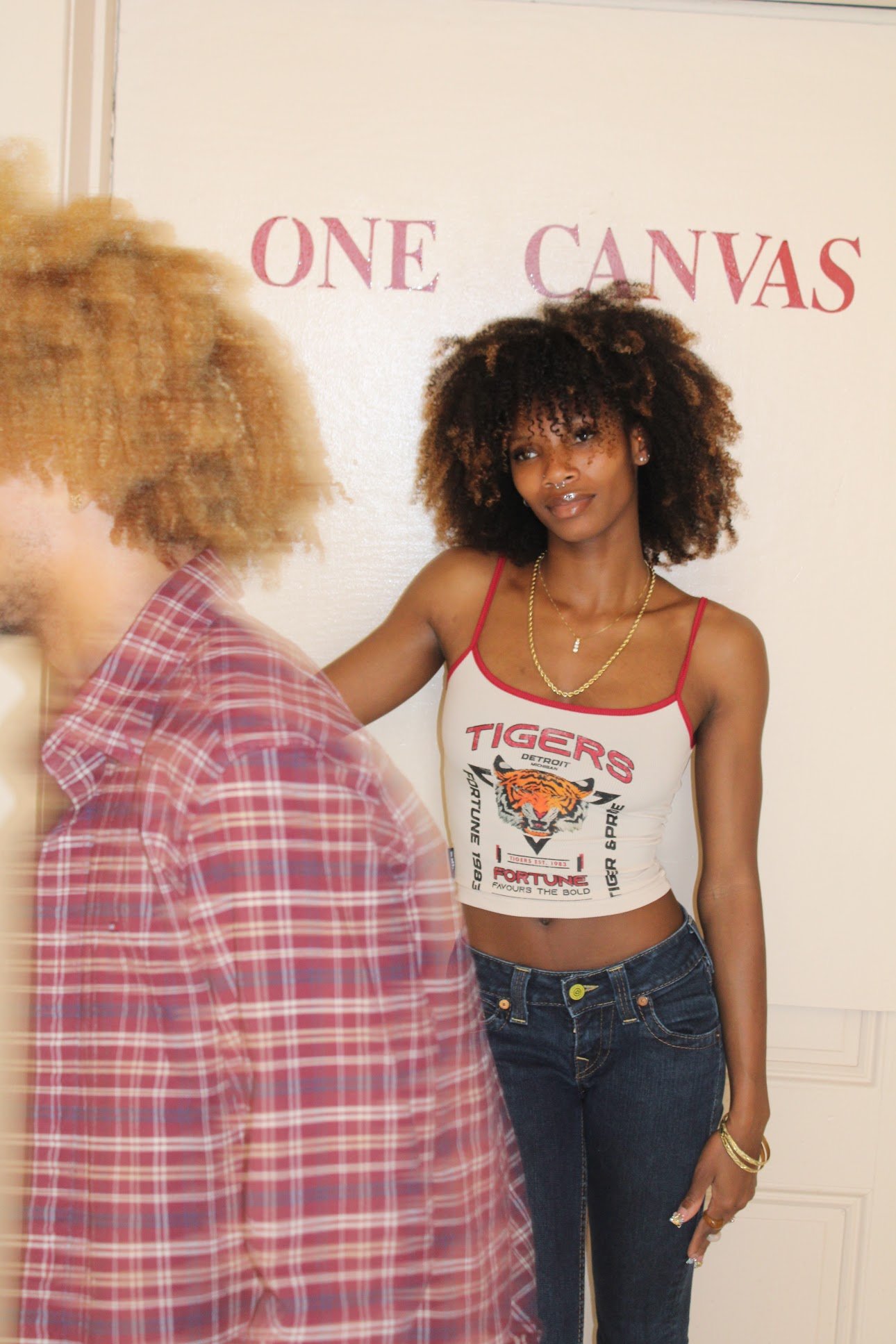 A woman with curly hair wearing a Detroit Tigers crop top and jeans, standing in front of a white wall with the words "ONE CANVAS" painted on it.