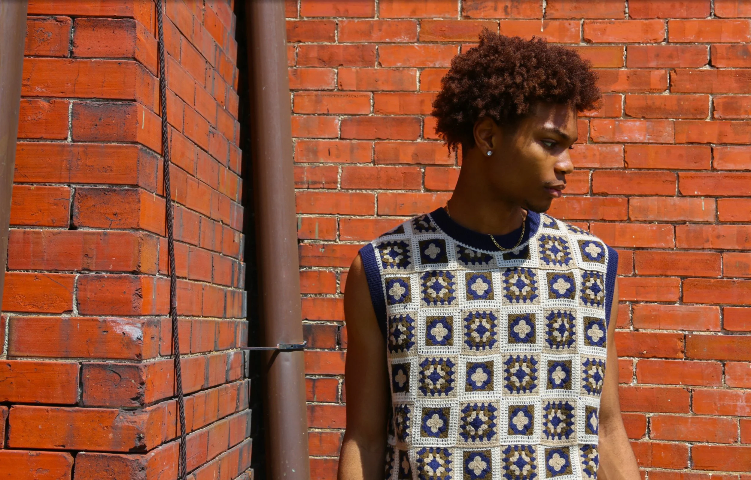 Young person with short curly hair wearing a sleeveless patterned top, standing next to a red brick wall and a brown pipe.