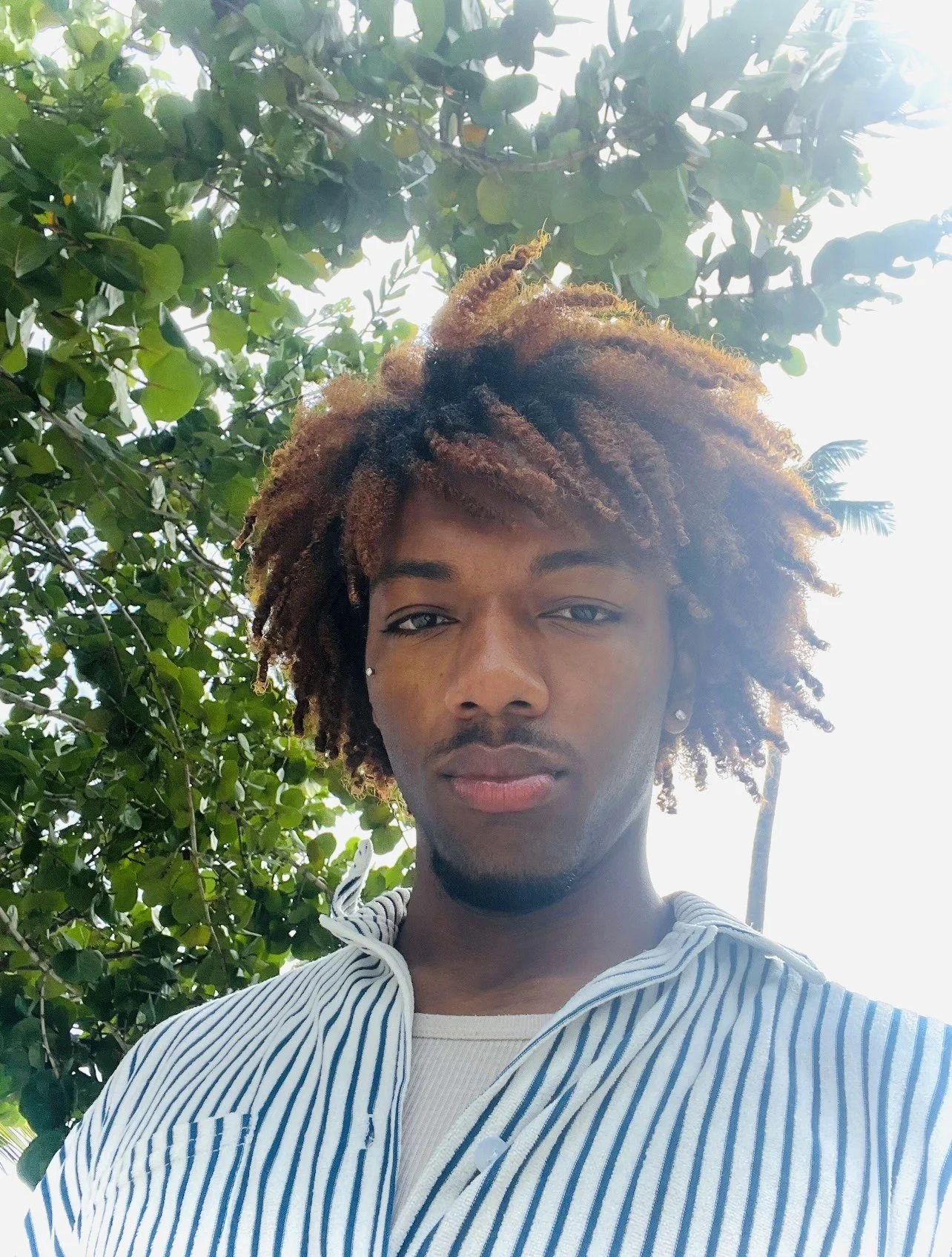 Young man with dreadlocks taking a selfie outdoors, with lush green trees and sunlight in the background.