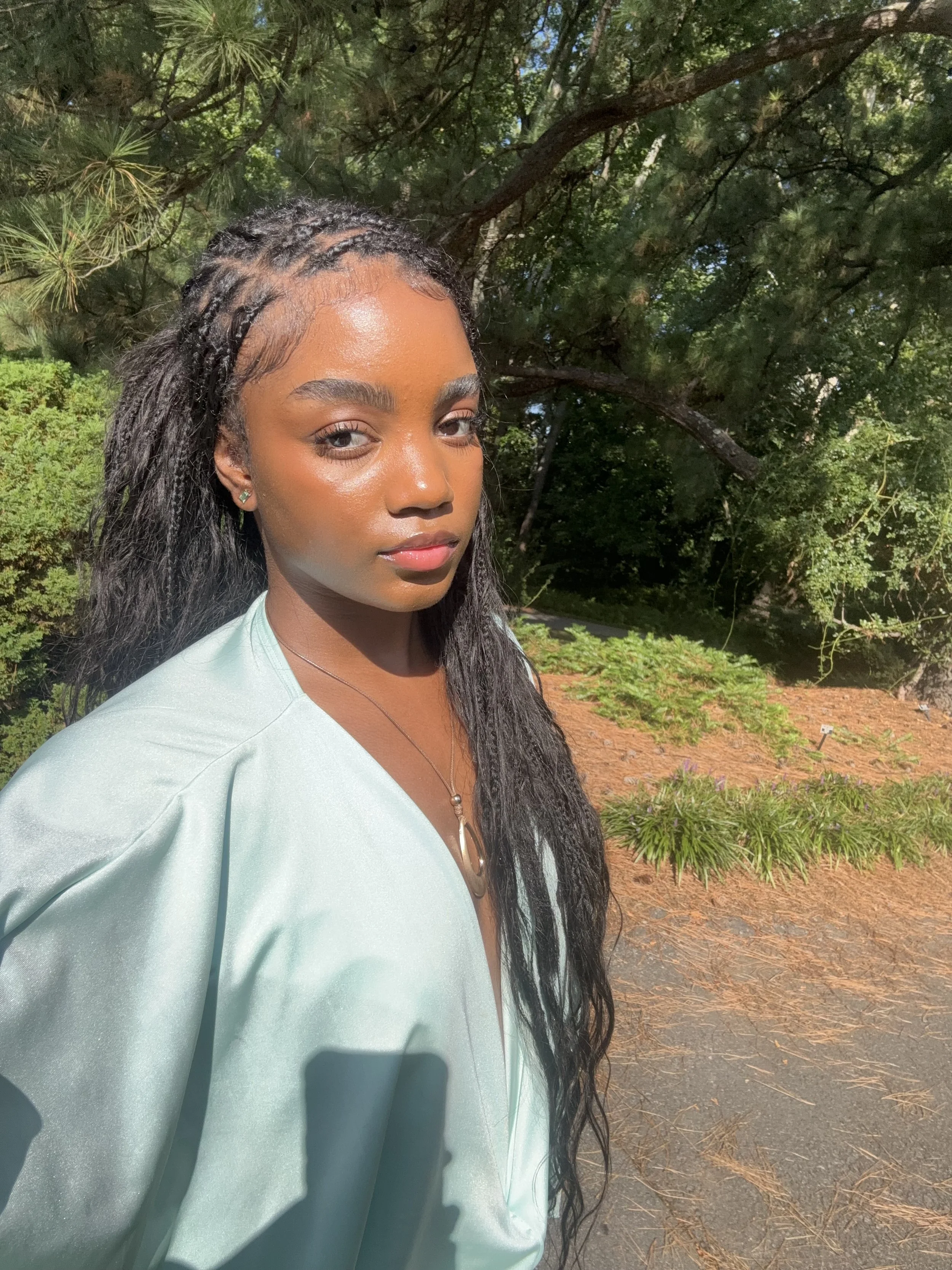A young woman with long, dark, braided hair standing outdoors in sunlight, wearing a light-colored top and a necklace, in front of green trees and plants.