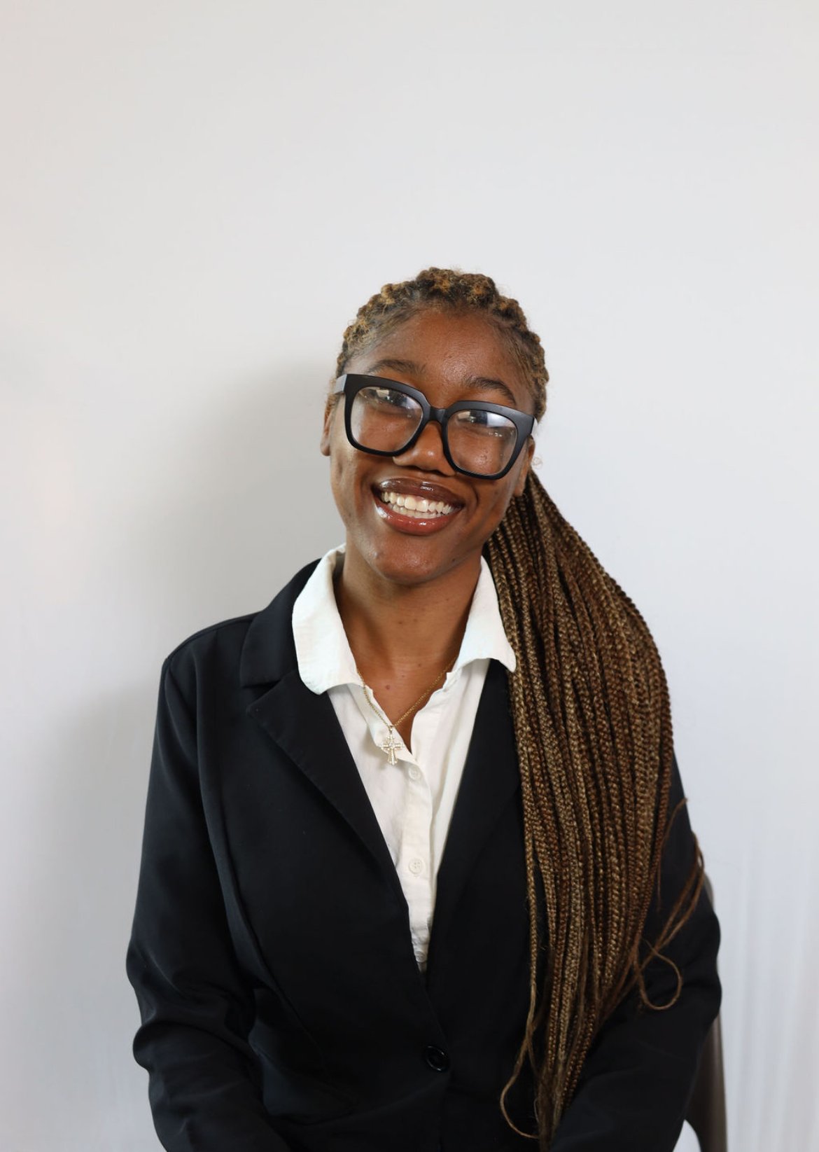 A smiling woman with dark skin, long braided hair, wearing large black glasses, a white collared shirt, and a black blazer, sitting against a plain white wall.