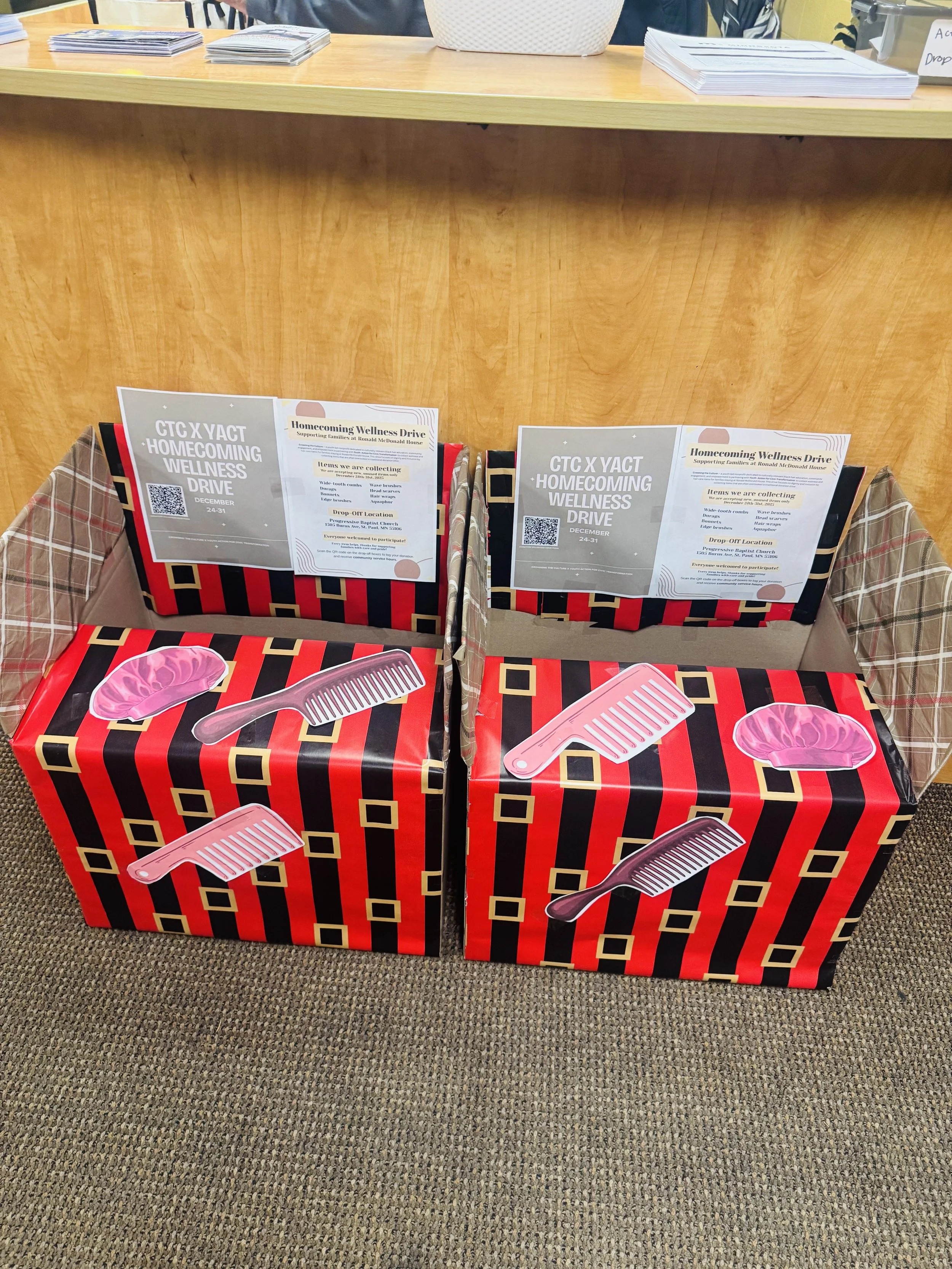Two donation boxes with red and black striped wrapping paper, each decorated with salon-themed stickers of combs and a hairbrush, placed on a carpeted floor in front of a wooden desk.