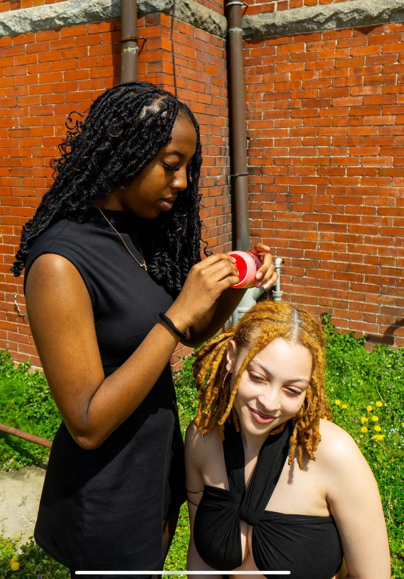 A woman with dark curly hair applying hair dye to a woman with blonde dreadlocks, sitting outdoors near a brick wall with green plants in the background.