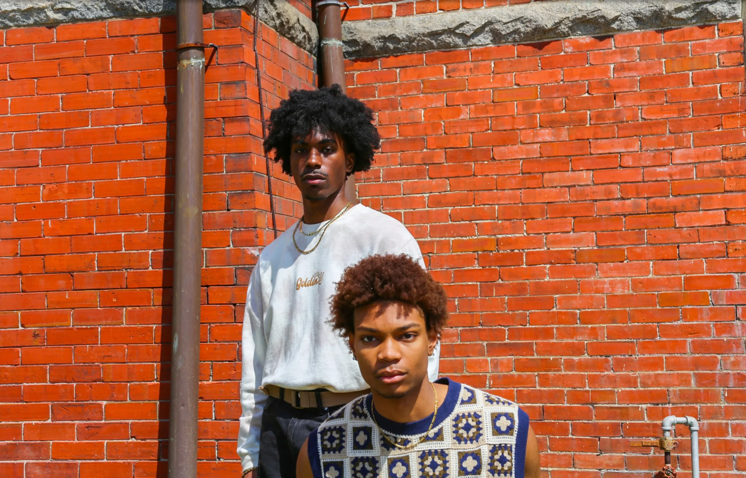 Two young men with curly hair standing in front of a red brick wall, one in a white sweatshirt and the other in a sleeveless patterned top.