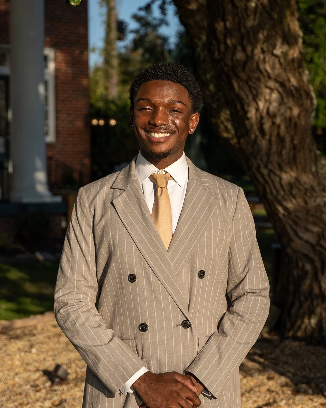 A young man in a beige pinstripe suit, white shirt, and gold tie standing outdoors next to a large tree, smiling at the camera.