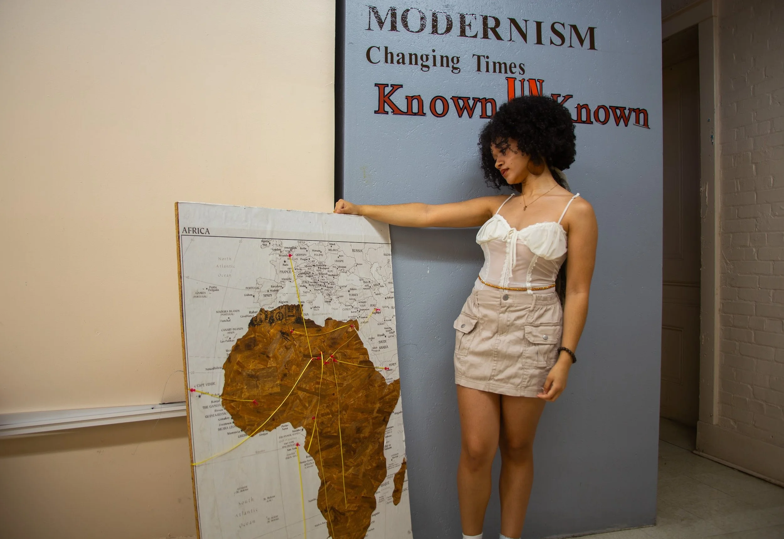 A woman with curly hair pointing at a map of Africa. The map is mounted on a board and shows lines connecting different parts of the continent. Behind her on the wall, there is a sign that reads 'MODERNISM Changing Times Known Unknown'.