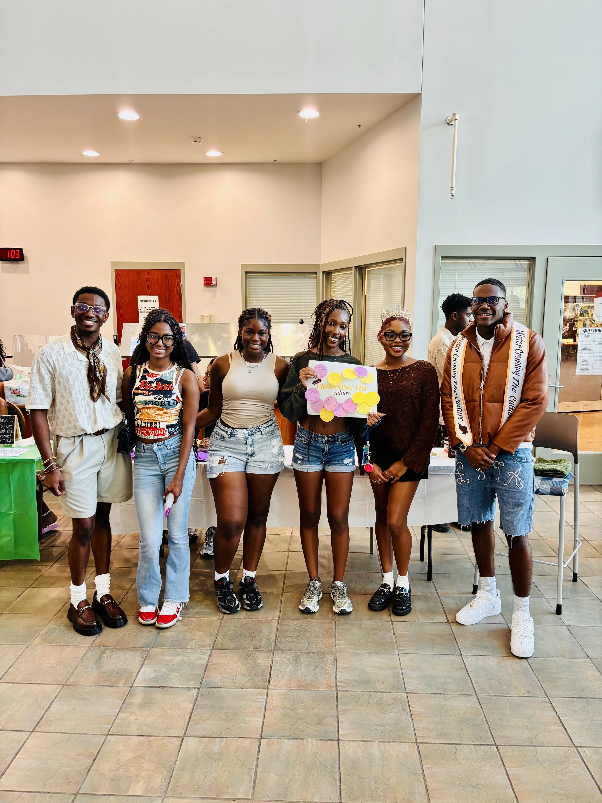 Group of six young people standing indoors in front of a table, smiling and posing for a photo. One person is holding a colorful sign that says 'Growing the Culture'.