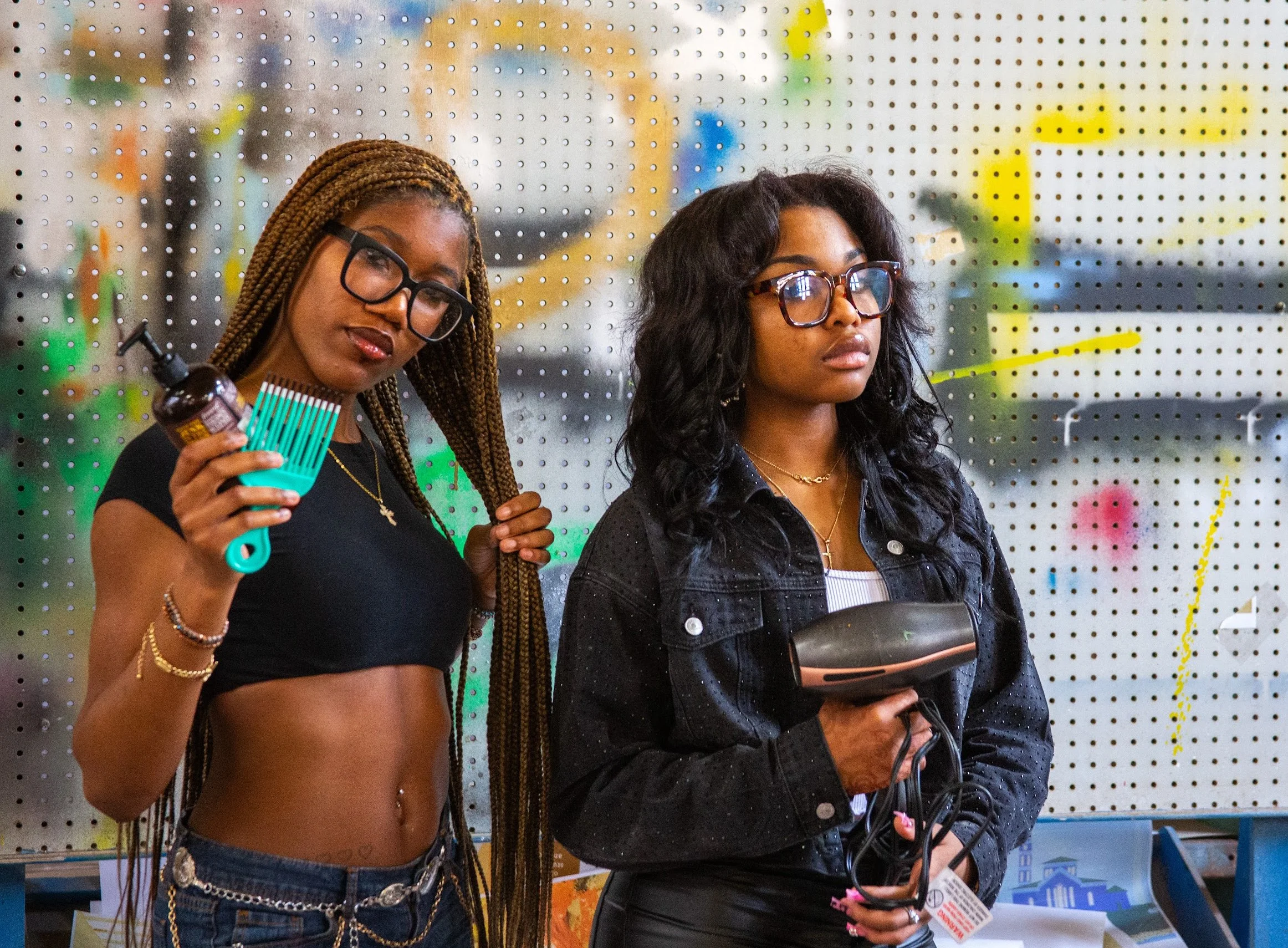 Two women shopping in a store, one holding a brush and the other holding a curling iron, standing in front of a pegboard display with various items.