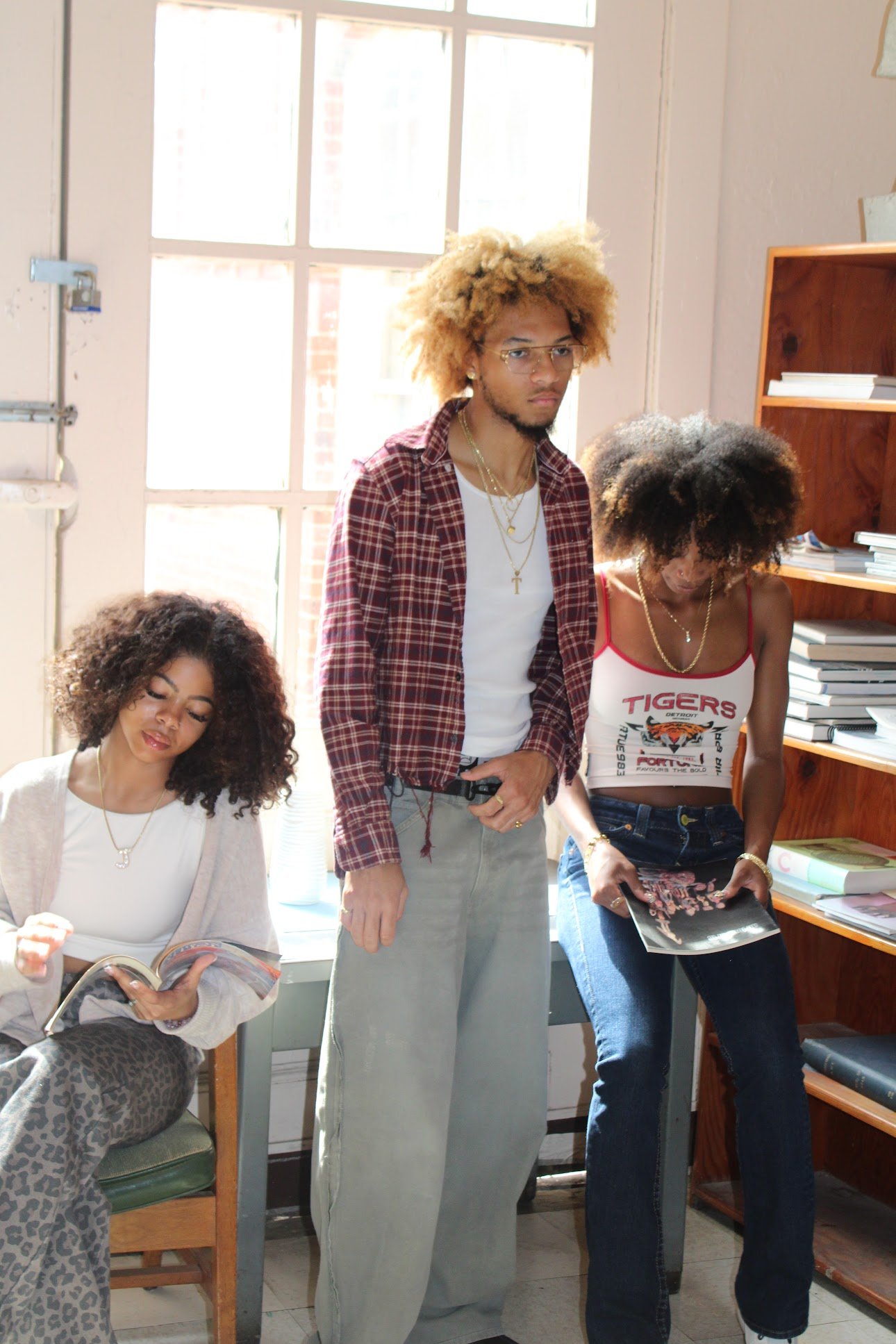 Three young women with curly hair in a room with sunlight, one reading a magazine, one standing with hands near pockets, and one looking at a book on a shelf.