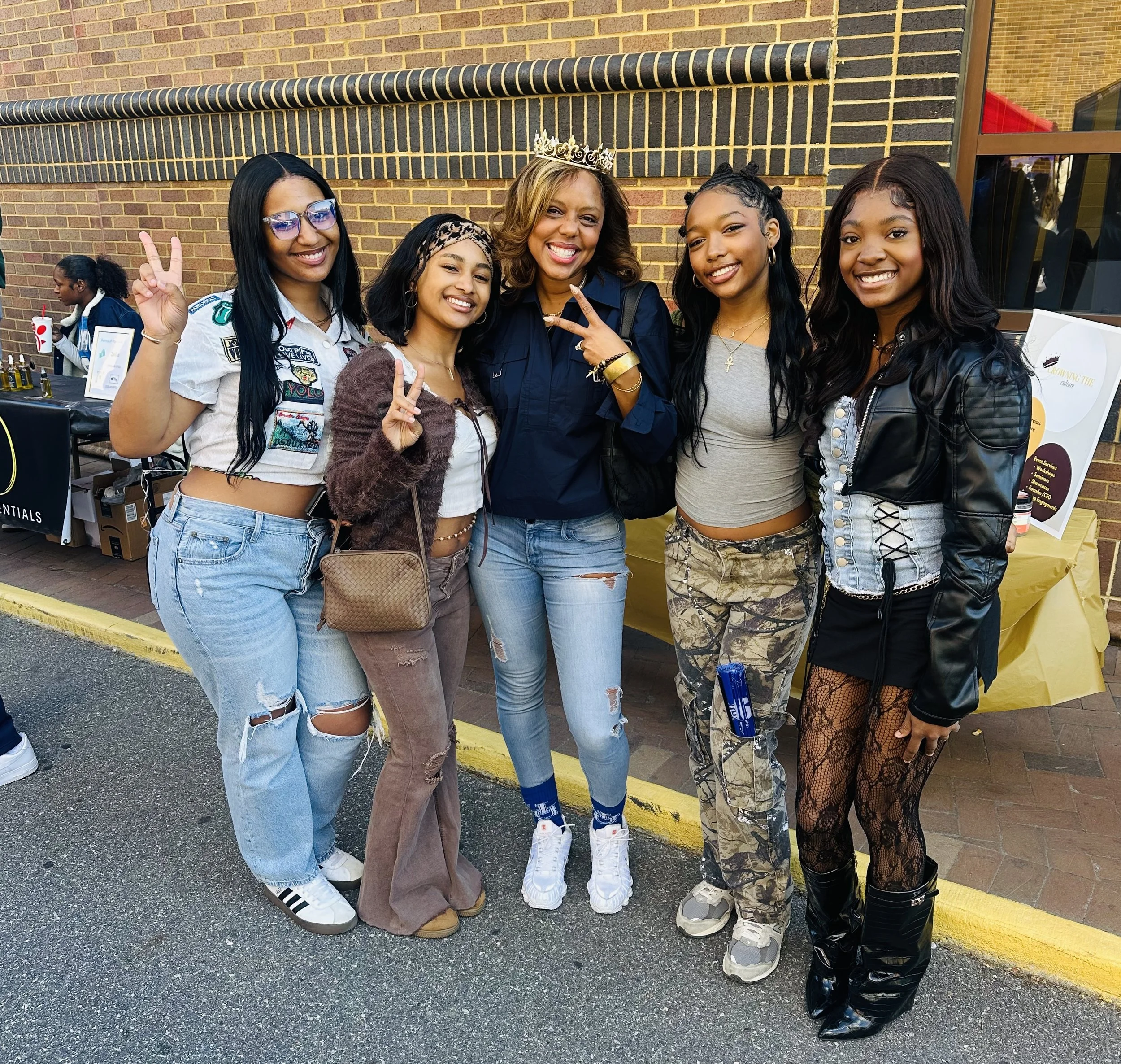 Group of five young women and one older woman smiling and making peace signs at an outdoor event, standing near a brick building, with tables and other people in the background.