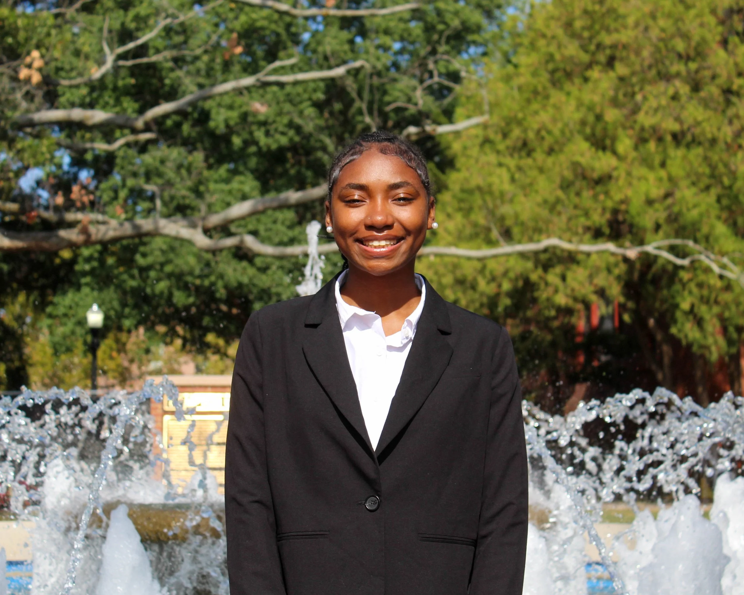 Young woman in black blazer and white shirt smiling standing in front of a fountain with trees in the background.