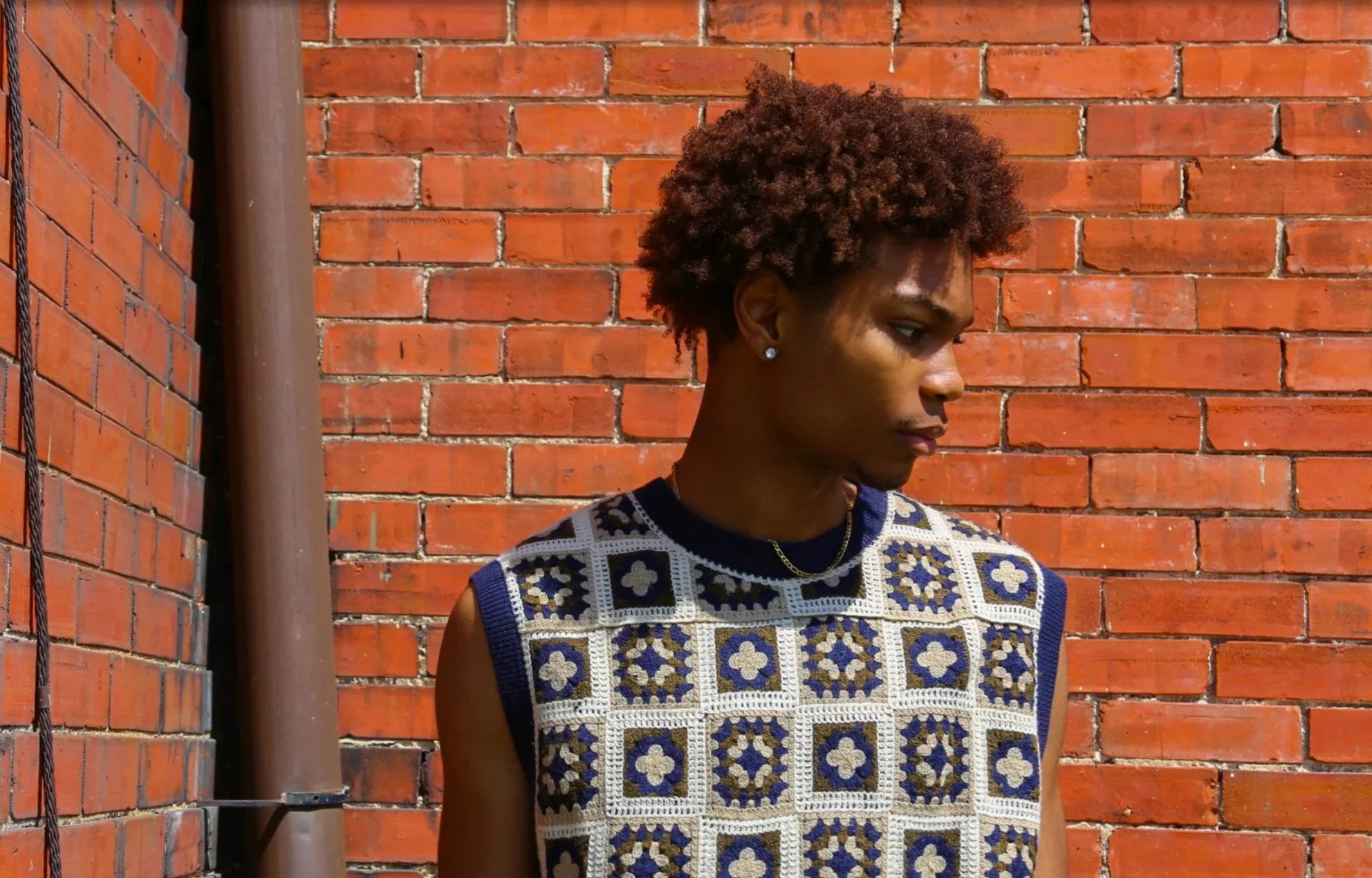 Young man with curly hair, wearing a sleeveless patterned vest, standing against a red brick wall.