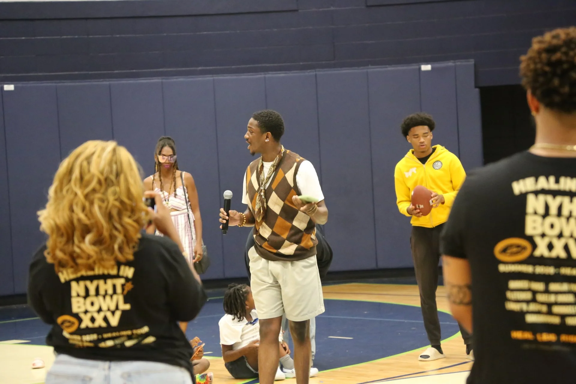 A group of young people in a gymnasium, some holding footballs, listening to a man with a microphone.