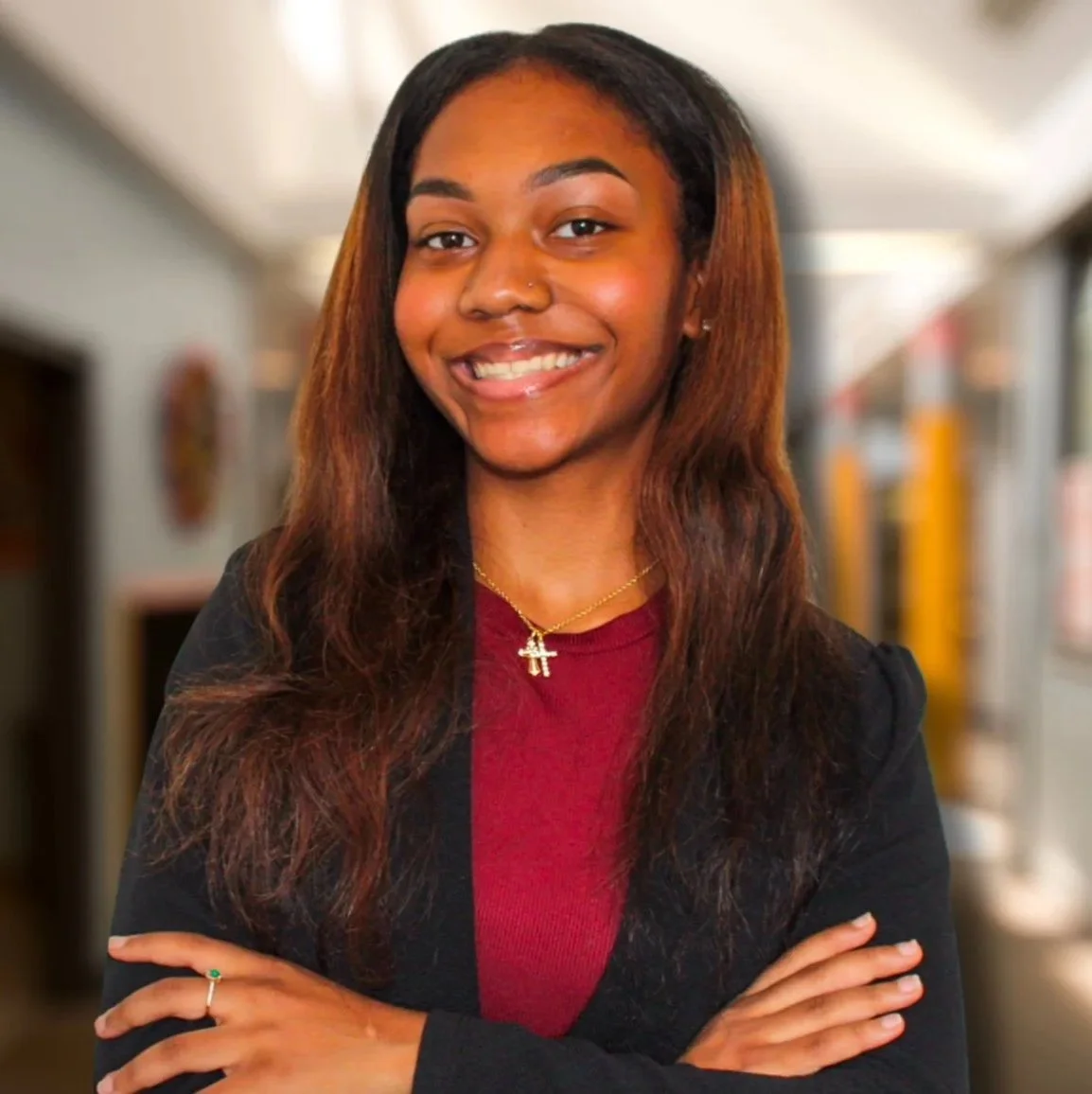 Portrait of a young African American woman with long hair smiling, wearing a red top and black blazer, standing in a blurred indoor setting.