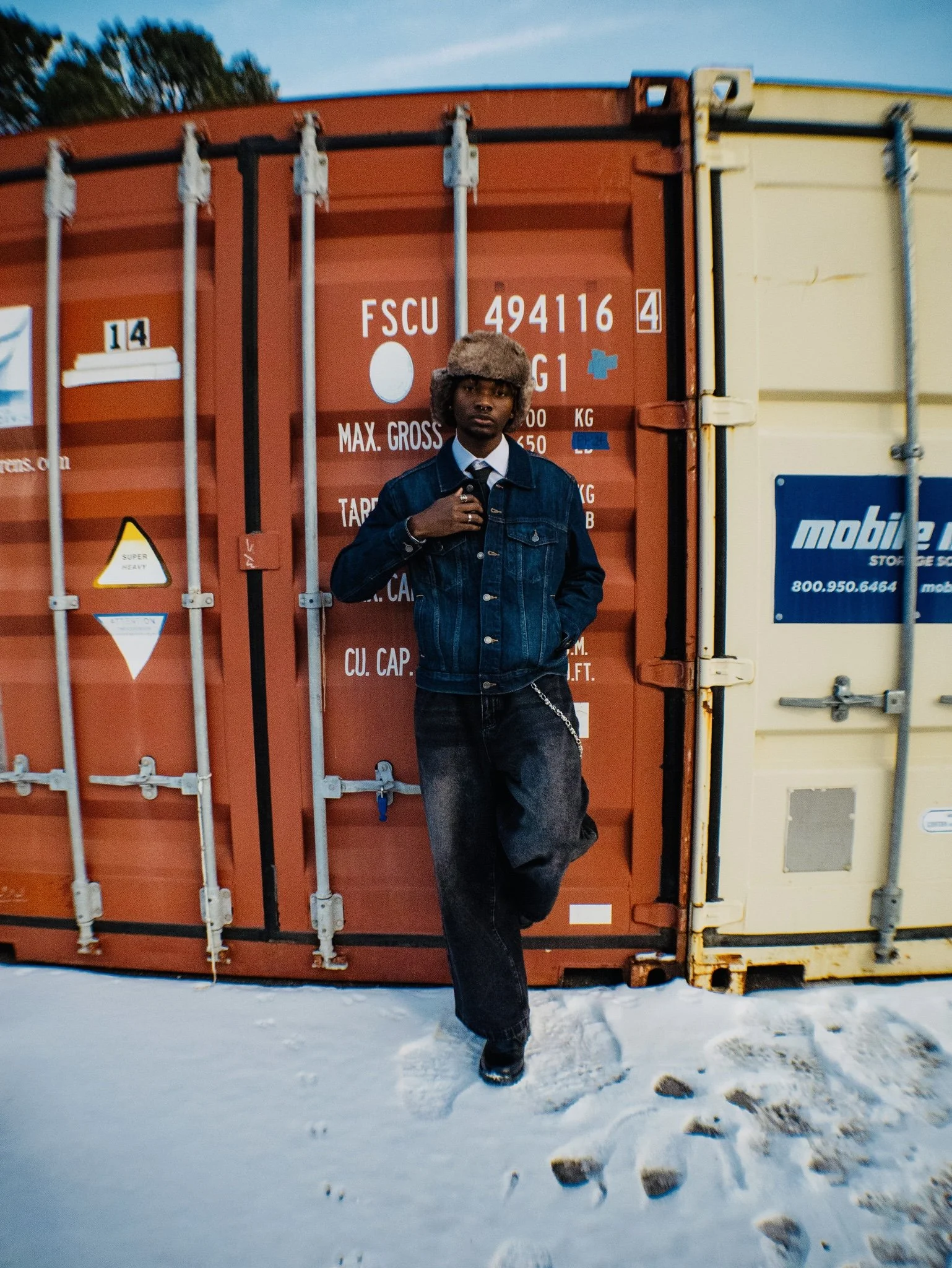 A person standing in front of stacked shipping containers on snow, wearing a fur hat, denim jacket, and jeans, with one leg crossed over the other.