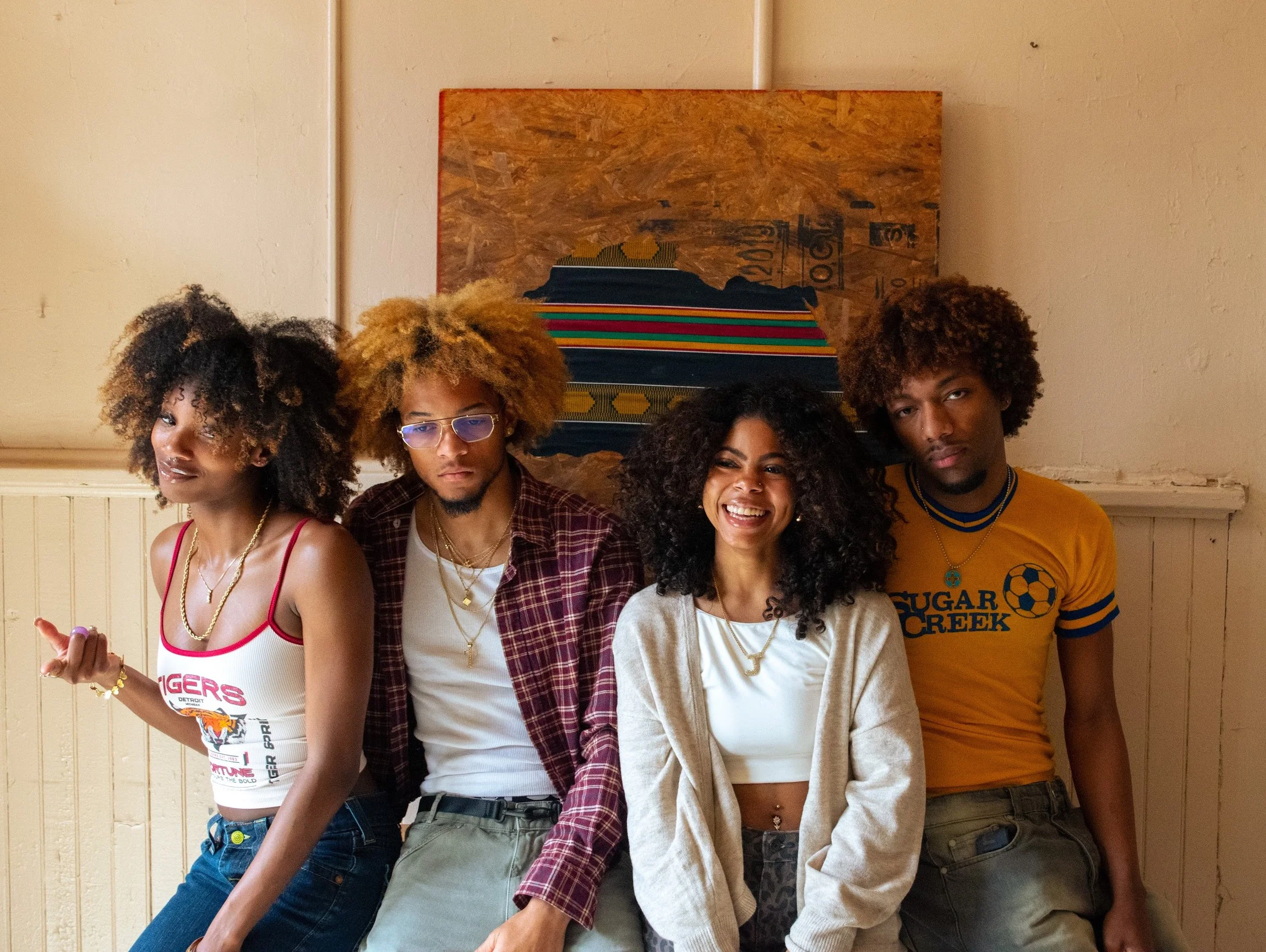 Four young adults with curly hair standing in front of a wall with an art piece, smiling and posing.