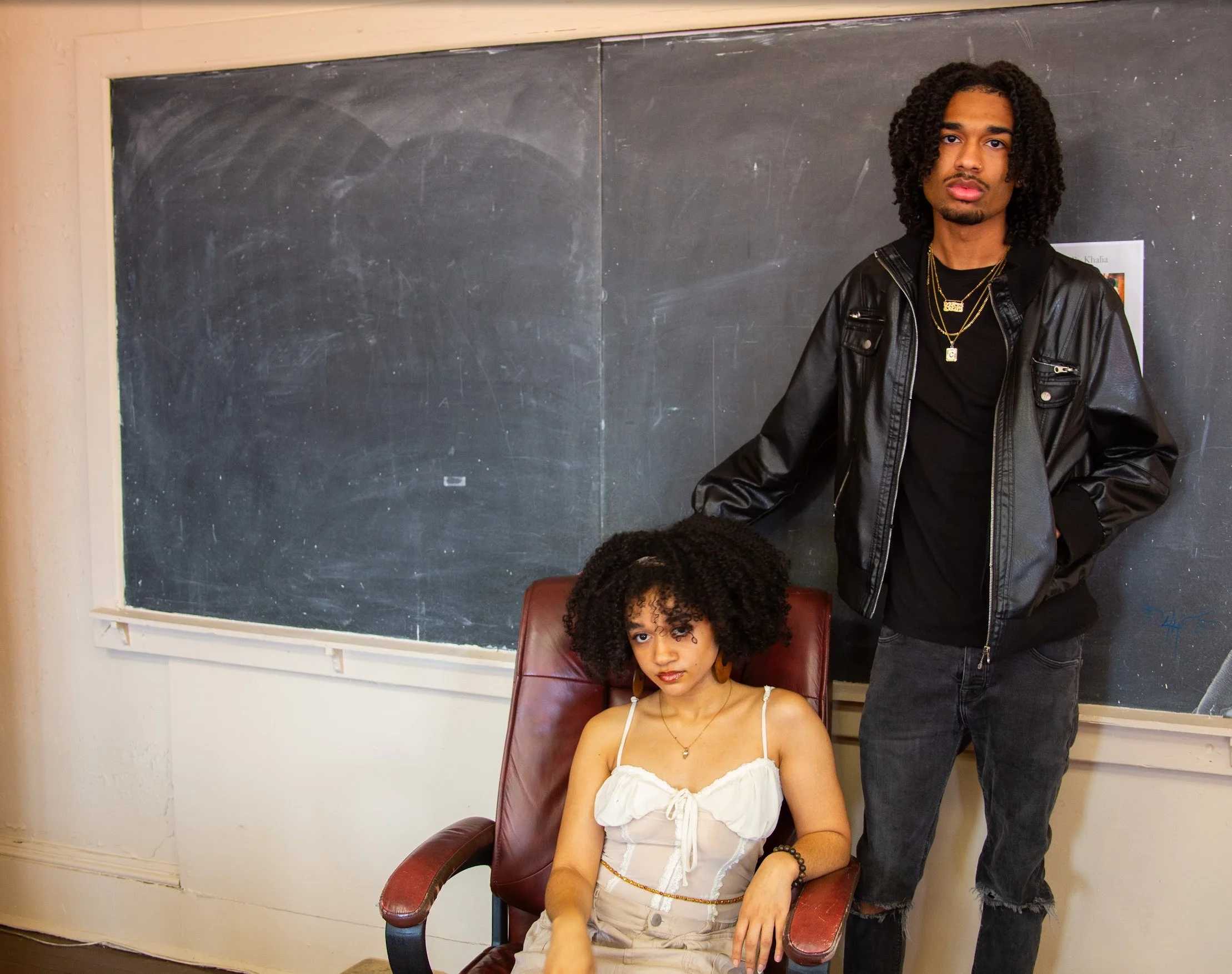 A young woman with curly hair, wearing a white crop top and beige pants, sitting on a red chair in front of a chalkboard. A young man with curly hair, wearing a black leather jacket, black t-shirt, and black jeans, stands beside her with one hand resting on the chair.