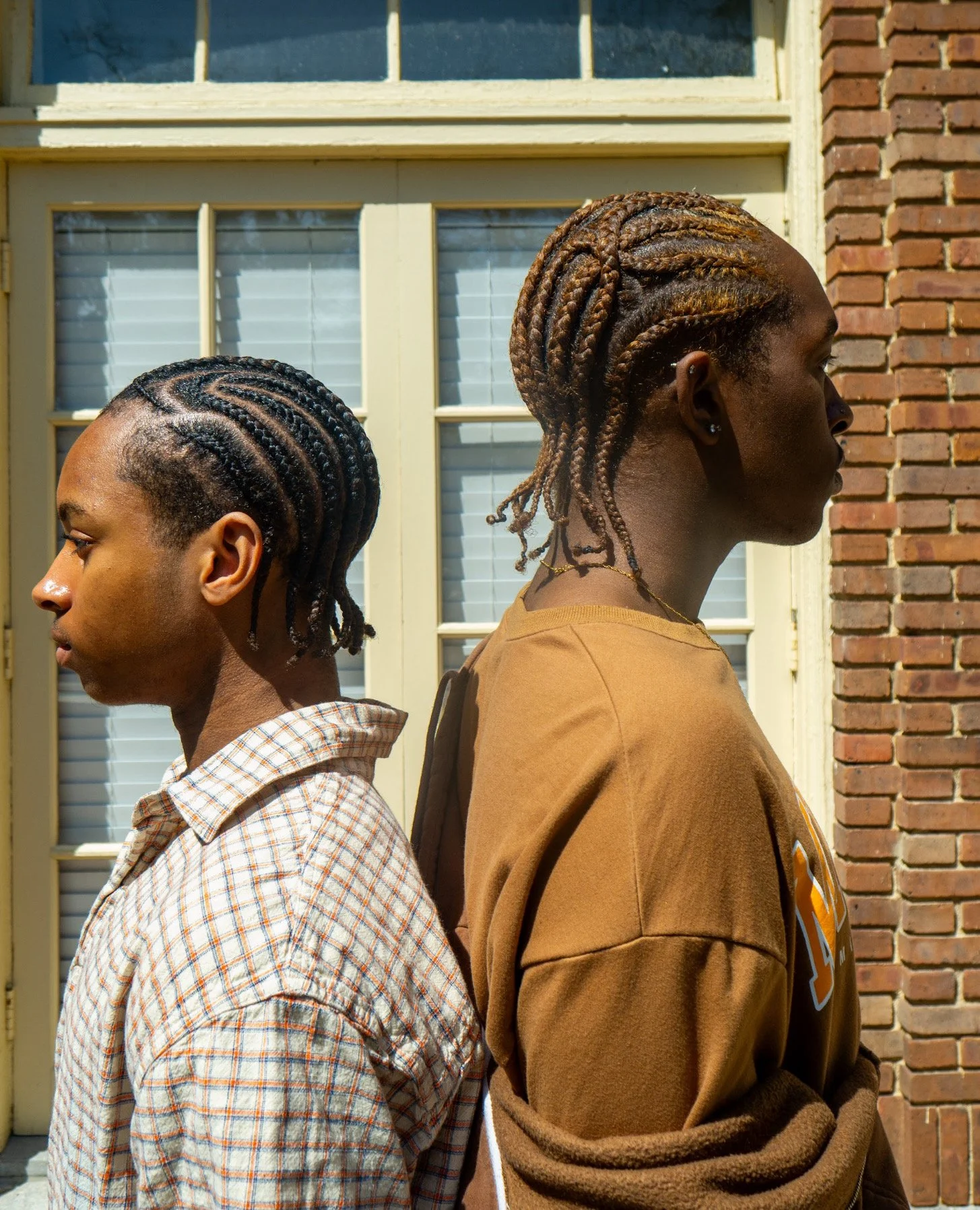Two young men with braided hairstyles standing back to back outdoors in front of a building with large windows and brick wall, facing opposite directions.