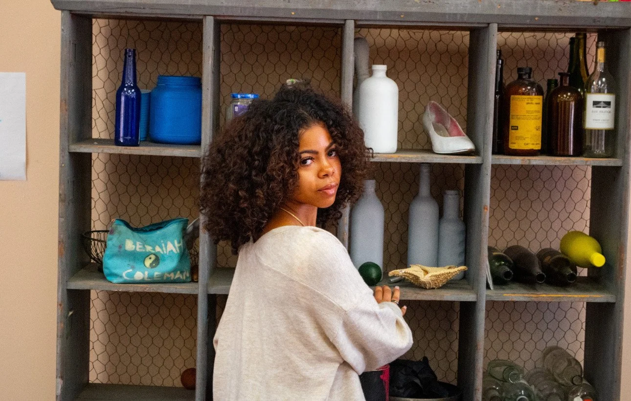 A woman with curly hair looking back over her shoulder in front of a wooden shelving unit with bottles, jars, and miscellaneous items.