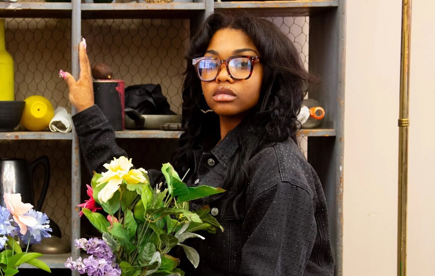 A woman with dark curly hair, glasses, and a black denim jacket stands next to a shelf, holding a bouquet of colorful flowers.