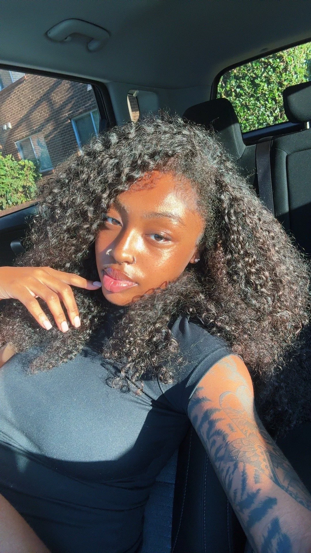 A young woman with curly hair and a nose ring sitting in a car, taking a selfie in sunlight with a tattoo on her arm and wearing a black top.
