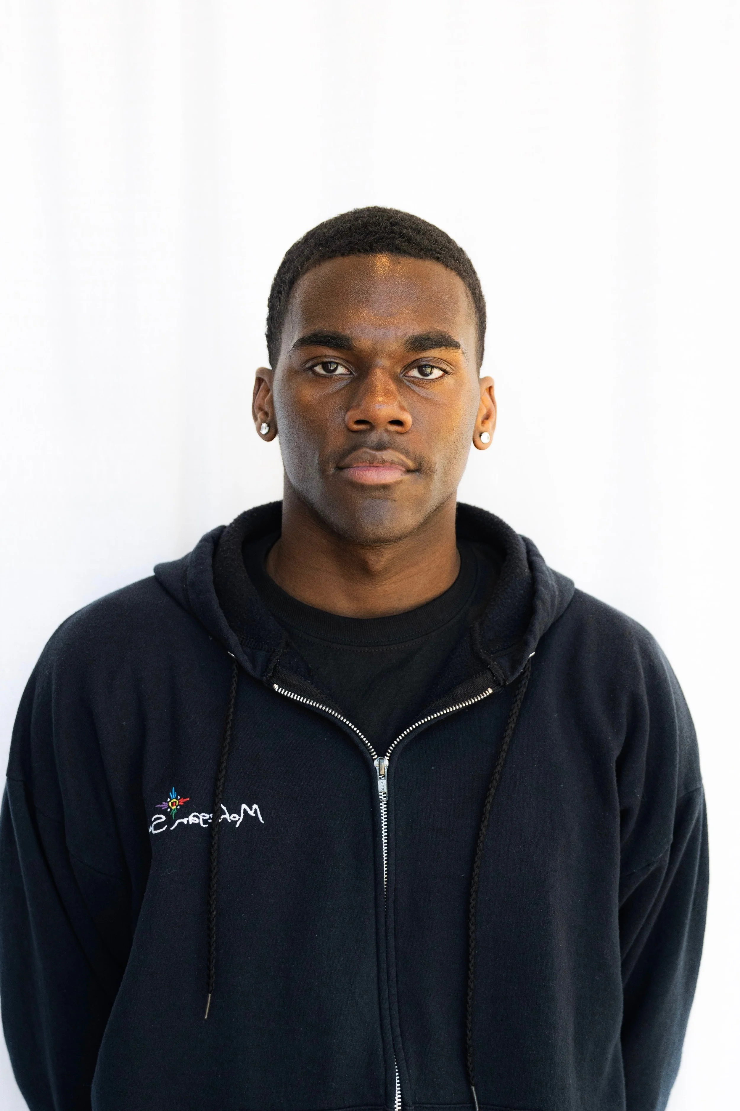 Portrait of a young African American man with short hair, wearing a black zip-up hoodie and earrings, standing against a plain white background. Black man with waves.