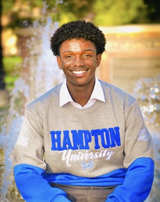 Young man smiling outdoors in front of a fountain, wearing a grey Hampton University sweatshirt with blue lettering.