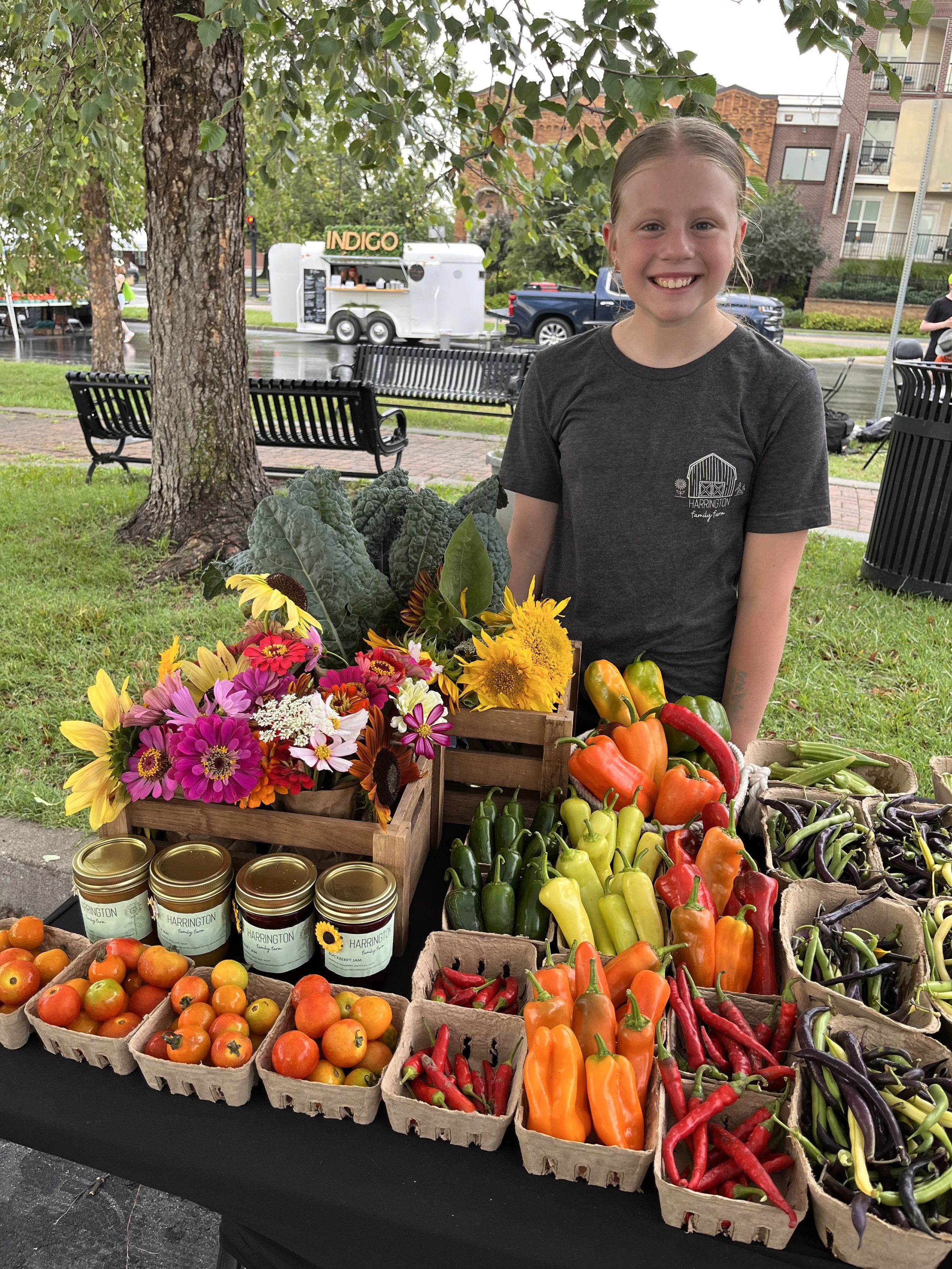 A girl with a big smile standing behind a table of fresh vegetables and flowers at an outdoor market.