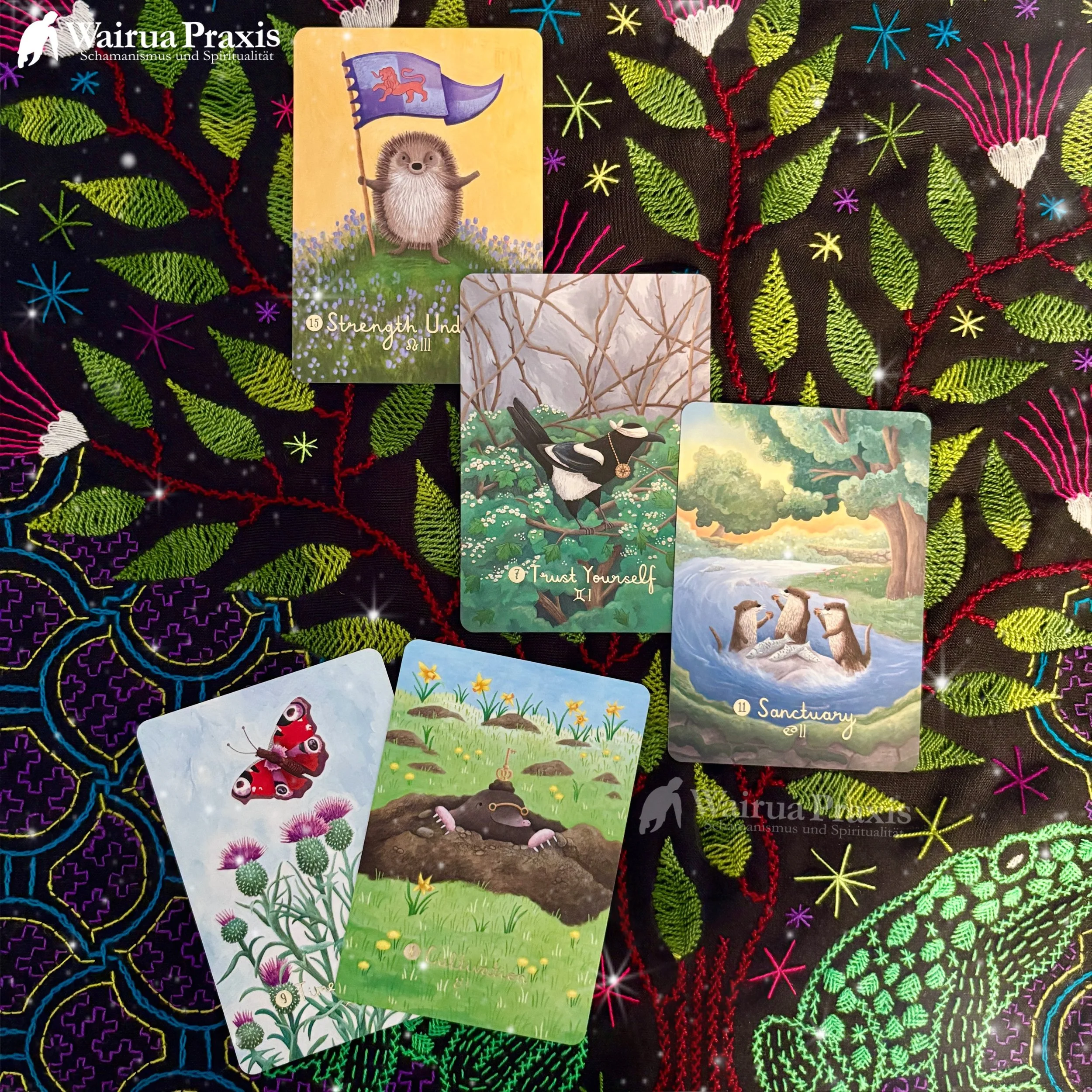 Mehrere Tarotkarten auf einem bunten, bestickten Stoff mit Pflanzen- und Blütendesign. Die Karten zeigen verschiedene Natur- und Tiermotive, darunter ein Igel mit einer Flagge, einen Spatz, Otter im Wasser, einen Schmetterling, eine Erdkröte und eine