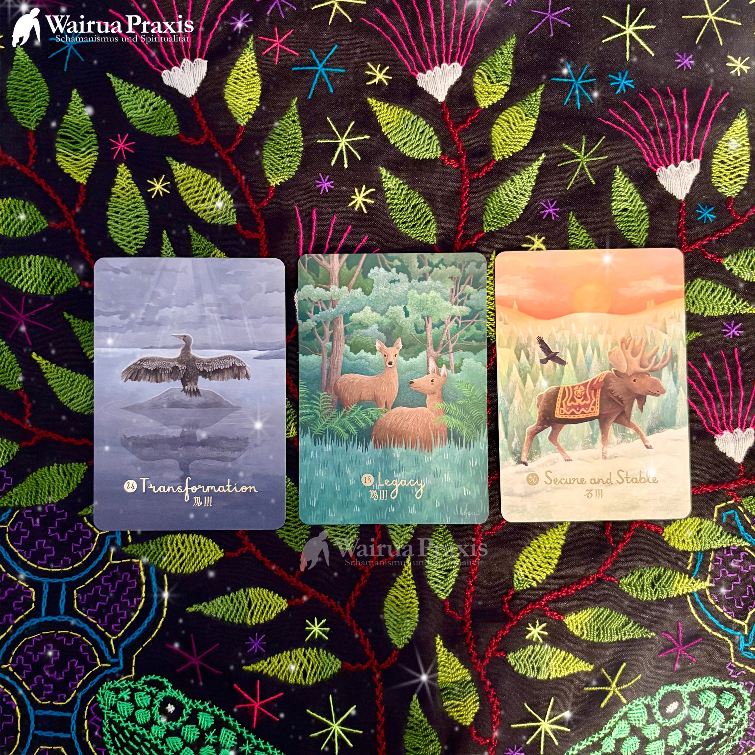 Drei Tarot-Karten auf einer schwarzen, bestickten Stoffdecke mit bunten, blattförmigen und sternförmigen Mustern. Die erste Karte zeigt einen schottischen Kranich mit dem Wort "Transformation". Die zweite Karte zeigt Rehe in einem Wald mit dem Wort "