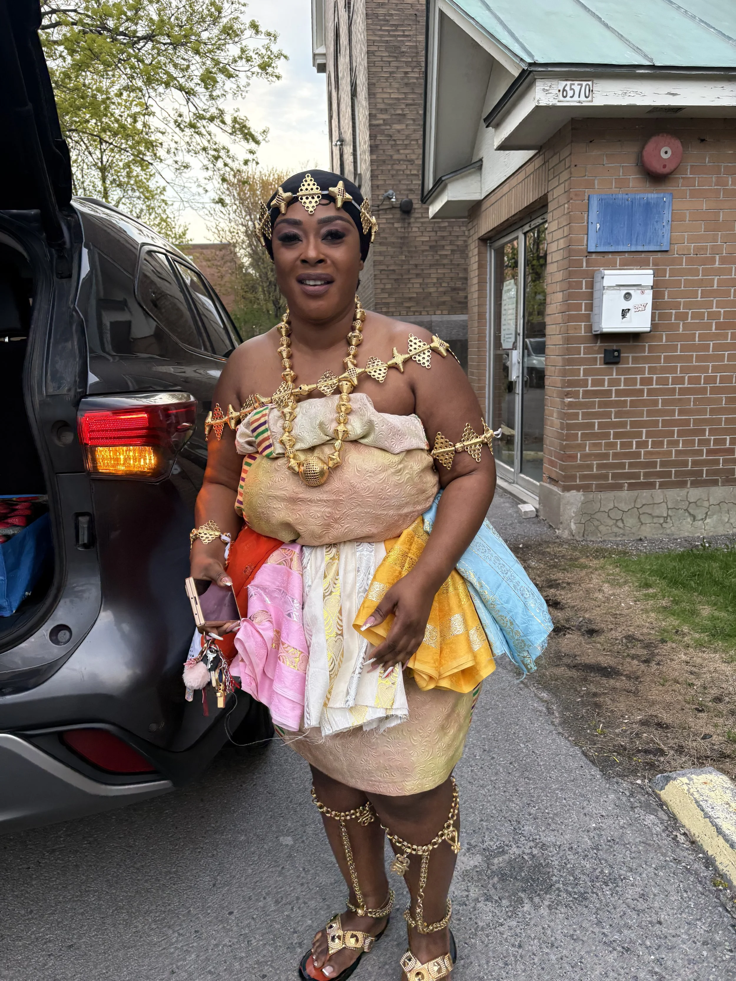 Woman dressed in elaborate gold jewelry and traditional attire standing outside near a car in front of a brick building with a blue sign and a mailbox.