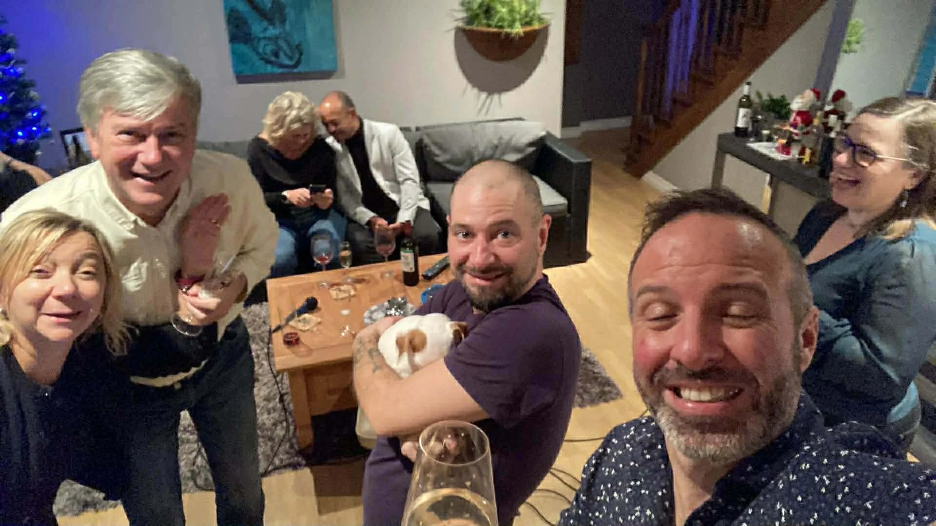A group of six adults smiling and gathered around a table at a party, with a Christmas tree in the background and Christmas decorations on a side table.