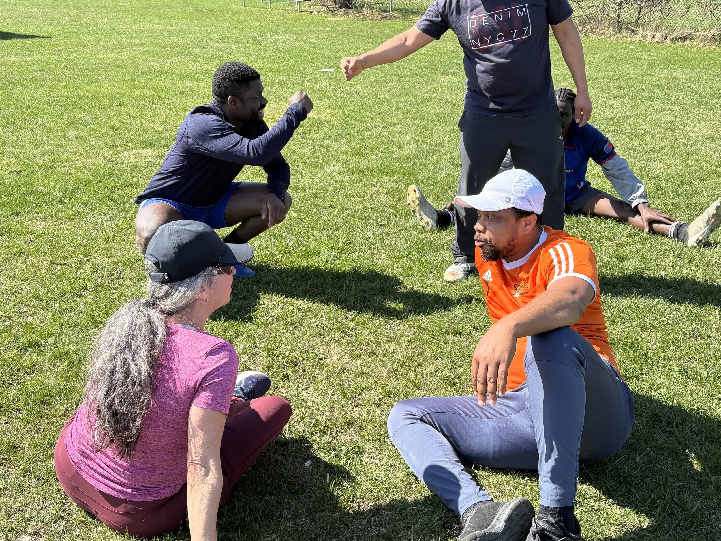 Four people sitting on the grass outdoors, engaged in a conversation or game, with a man standing and interacting with a woman in a pink shirt.