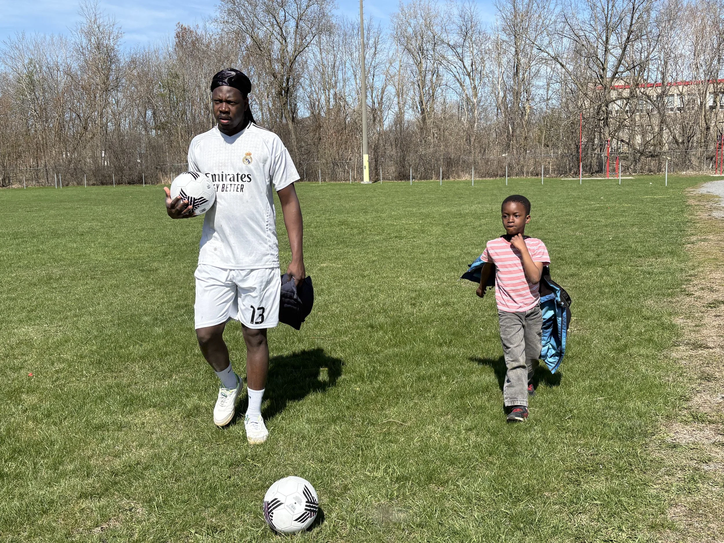 A man in a white soccer uniform holding a soccer ball walking on a grassy field, with a young boy carrying a backpack walking beside him. The background has leafless trees and a building, with a clear blue sky.