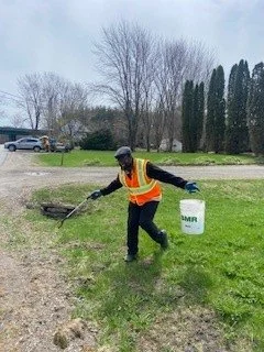 A person wearing a helmet, yellow safety vest, and black clothing, using a tool to remove weeds from a grassy area near a sidewalk, with trees and parked cars in the background.